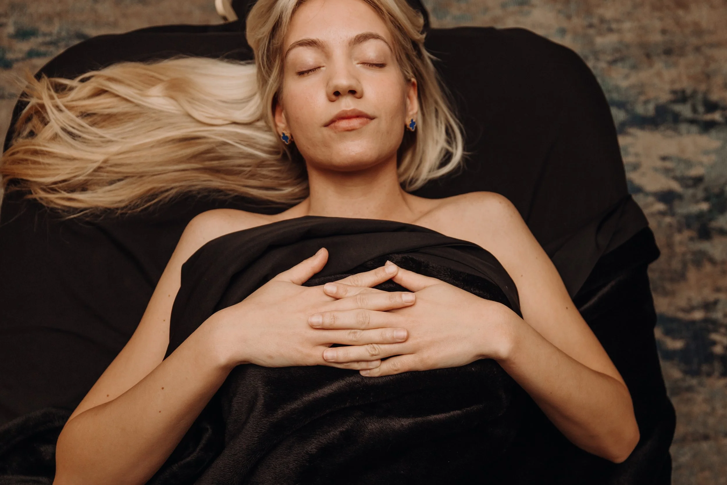 A woman with blonde hair and earrings is lying down with her eyes closed, covered with black fabric, on a dark surface.