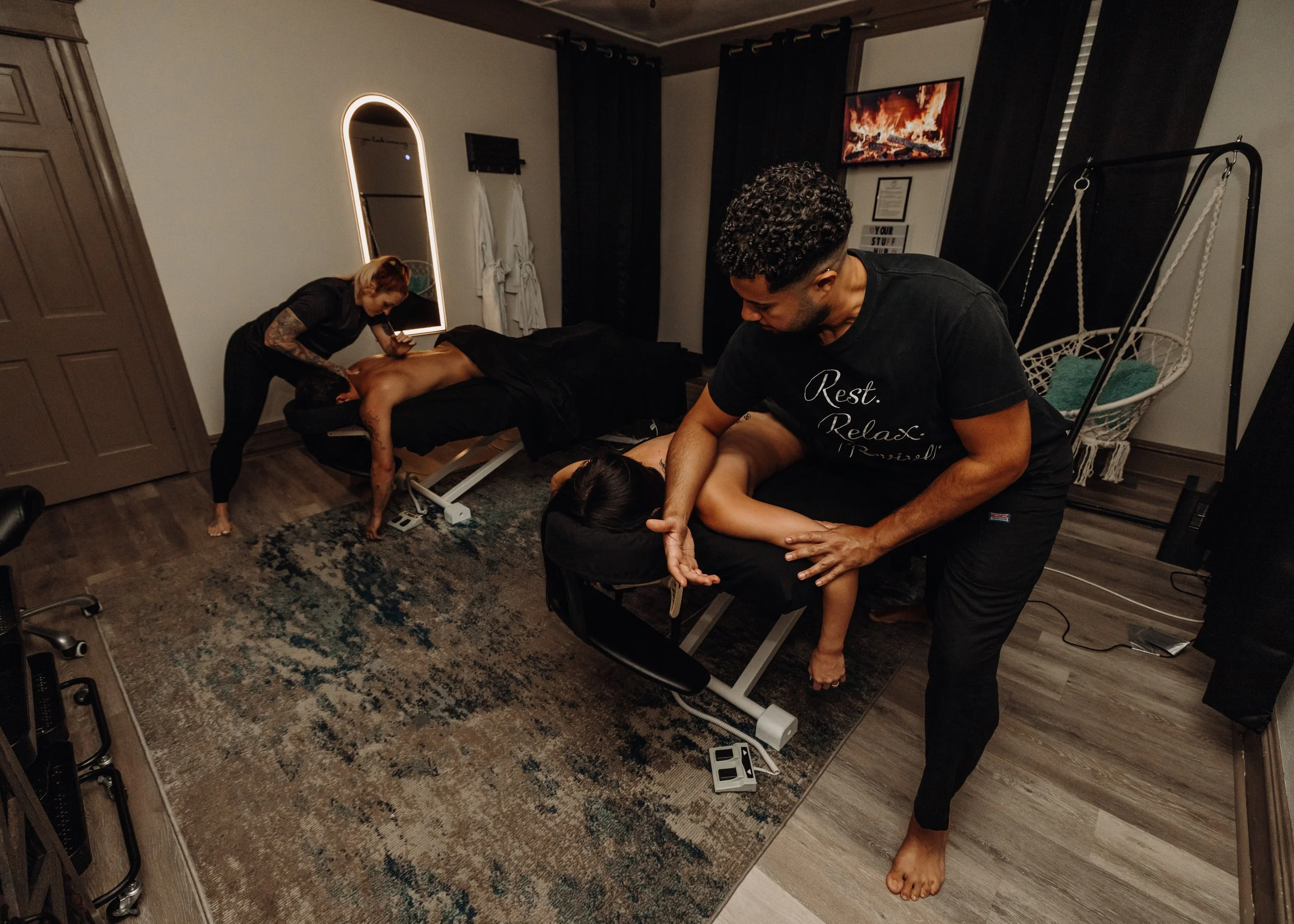 People receiving massages in a cozy room with dark curtains and a TV showing flames.