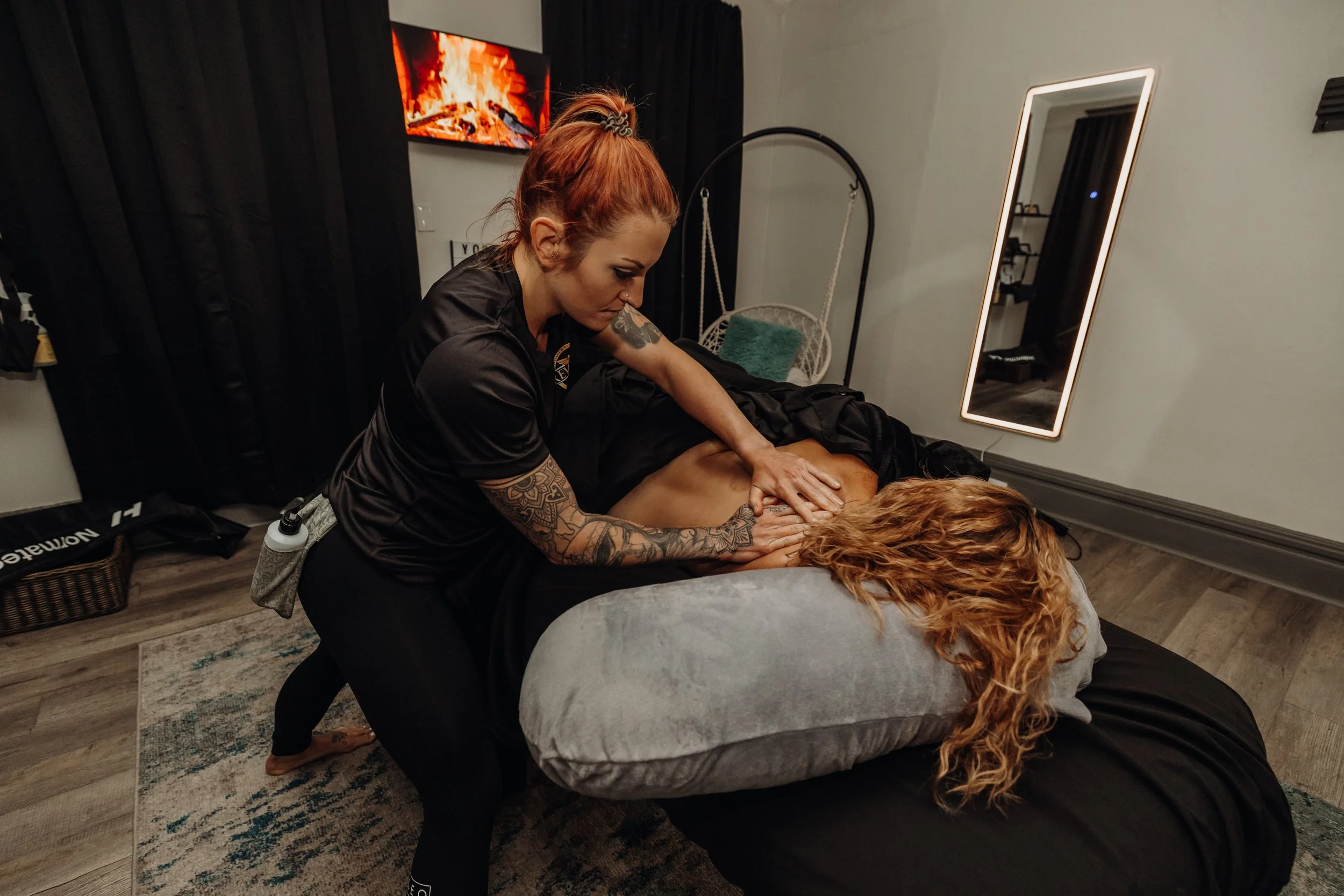 A woman with tattoos on her arms giving a massage to a person with long curly red hair lying face down on a massage table in a dimly lit room with a mirror, black curtains, and a fireplace TV.