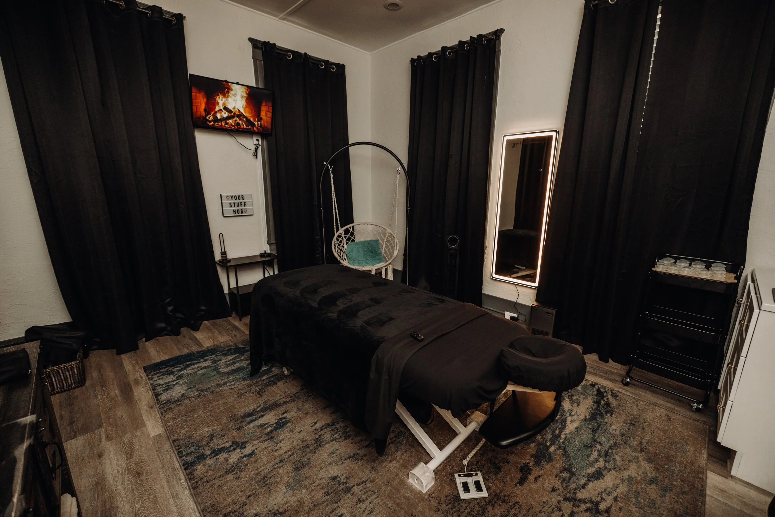 A cozy bedroom with black curtains, a wall-mounted TV showing a fireplace, a white hanging chair with a teal cushion, a black bed, a mirror with backlight, a small table with cups, and a wood floor with a colorful rug.
