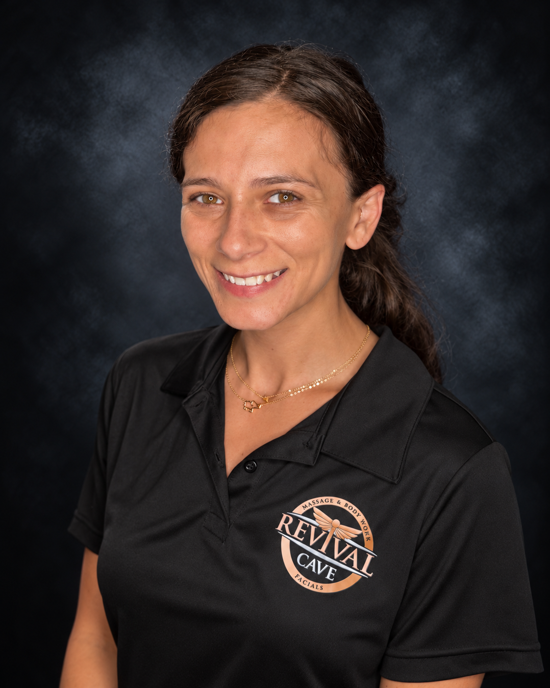 A woman with brown hair and hazel eyes smiling, wearing a black uniform with a logo for a massage and body work business over the chest, and a gold necklace.