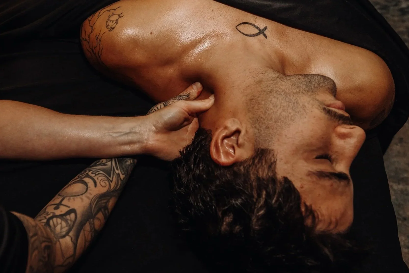A shirtless man with tattoos resting on a black surface, with a woman touching his face gently