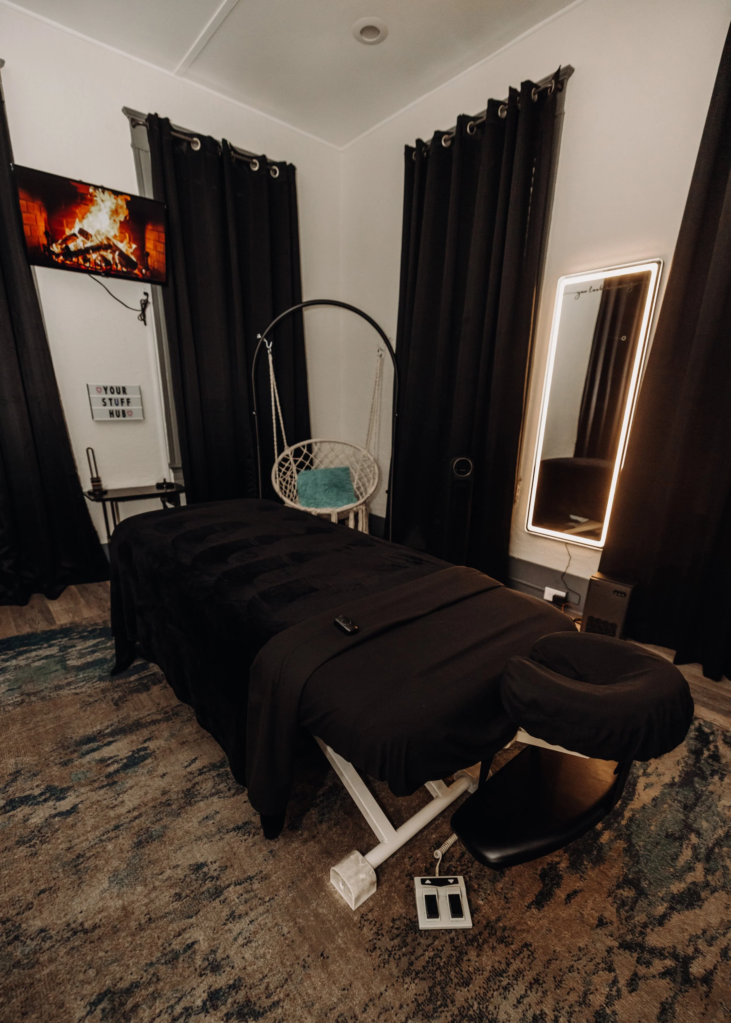 A bedroom with a black bedspread, black curtains, a full-length mirror with illumination, a hanging swing chair with a teal pillow, a television showing a fireplace, a small sign on the wall reading 'Your Stuff Hub,' a black chair, and a patterned carpet.
