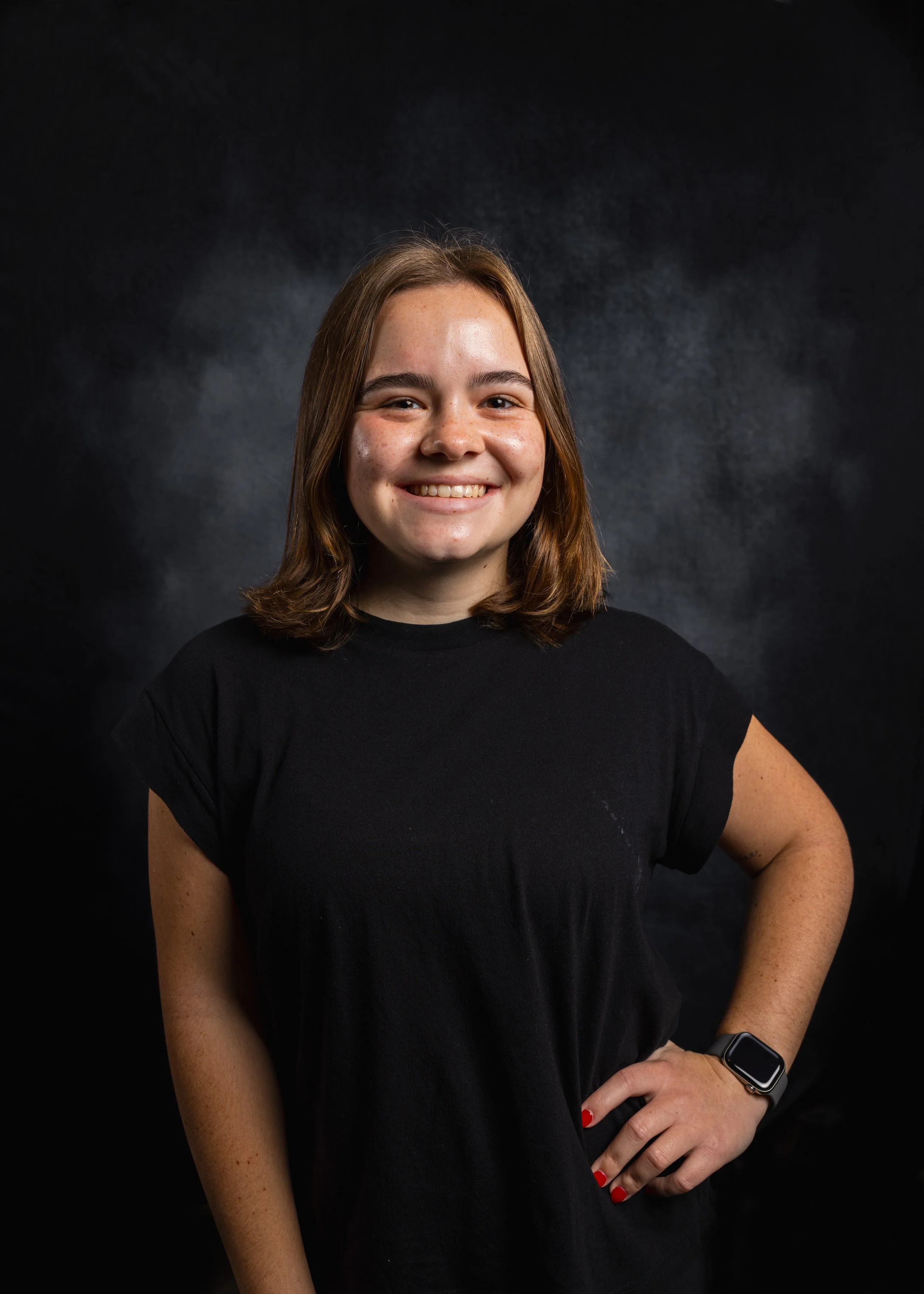 A young woman with shoulder-length brown hair and freckles, smiling and wearing a black t-shirt and a smartwatch on her left wrist, is standing against a dark background.