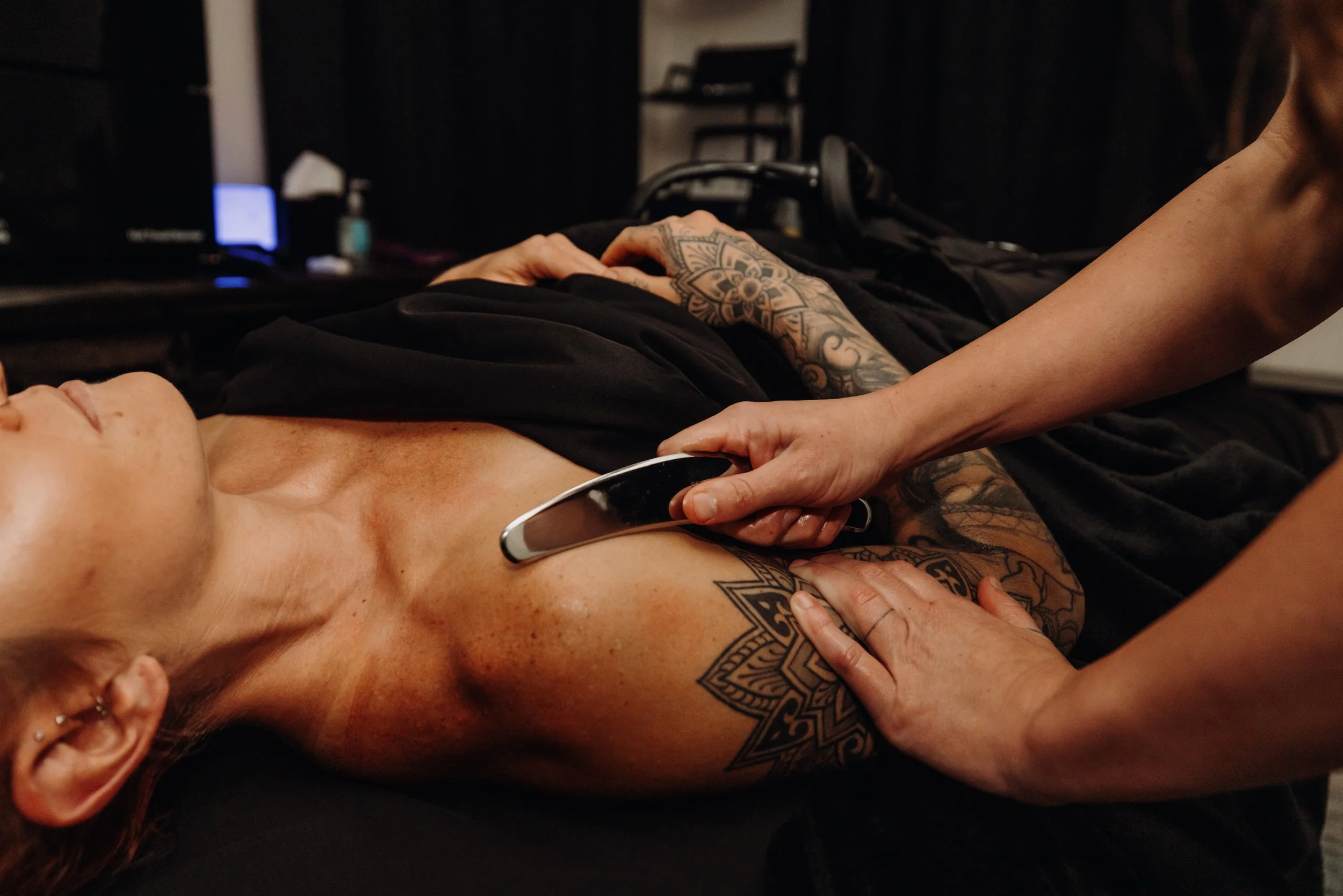 Close-up of a person with tattoos lying down and another person performing a treatment or therapy on them, with a dark background and some equipment visible.