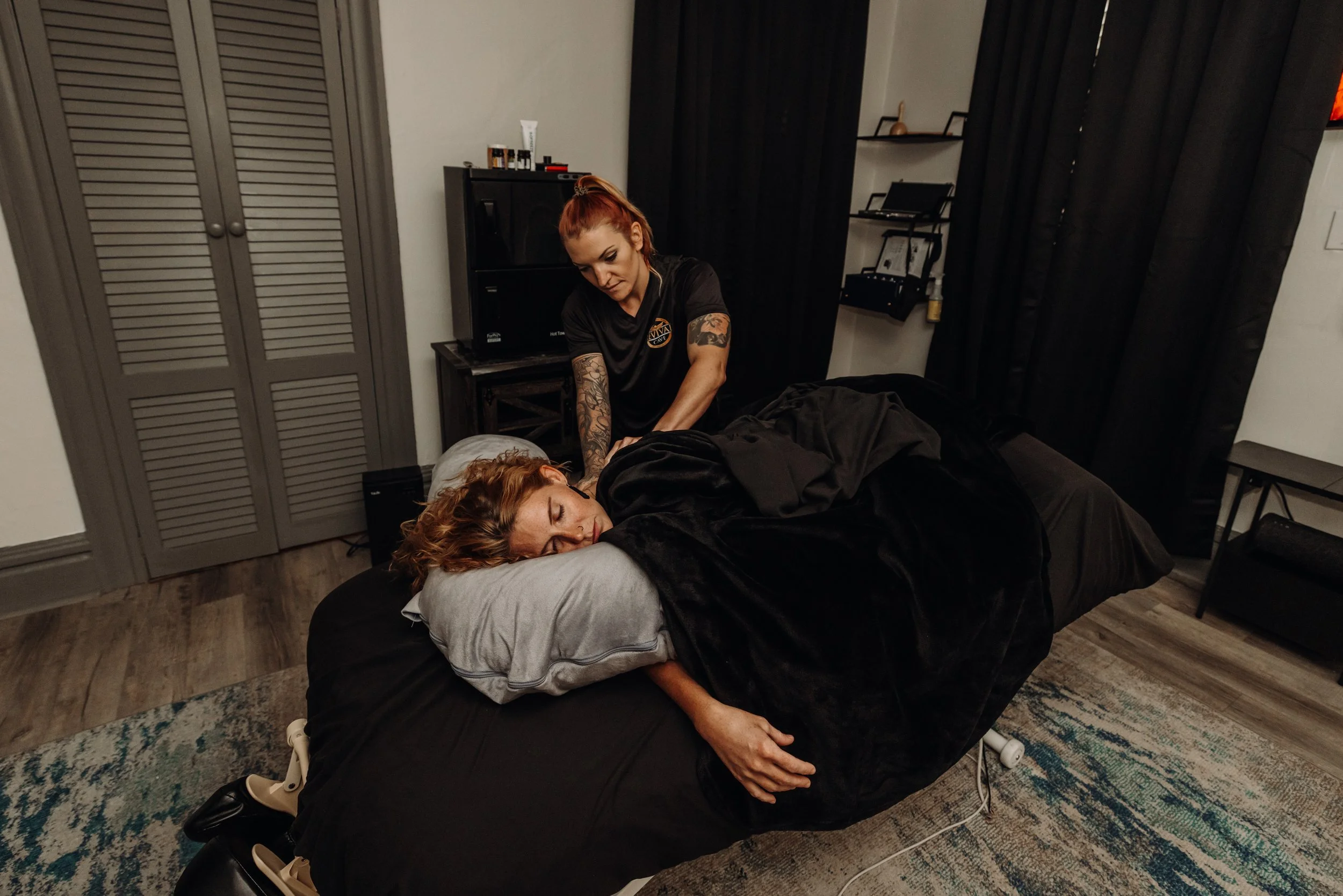 Woman with tattoos giving a massage or adjusting a person lying face down on a massage table in a dimly lit room.