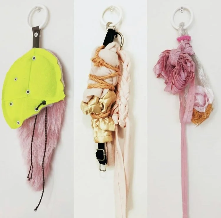 Three pink and green soft sculptures hang on a beige wall.