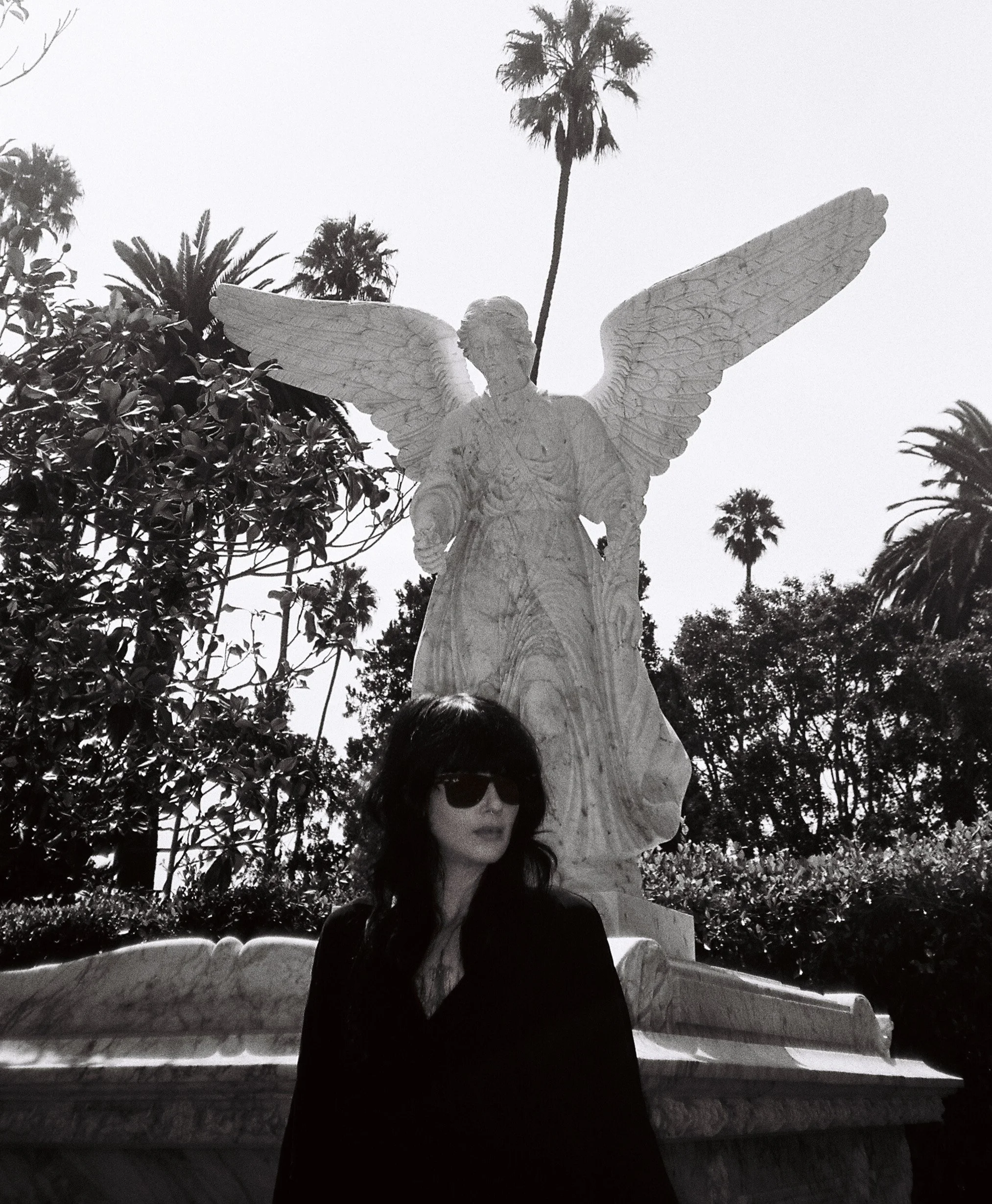 The writer Violetta Leigh stands in front of an angel statue in LA's Hollywood Forever cemetery in a black dress and sunglasses.