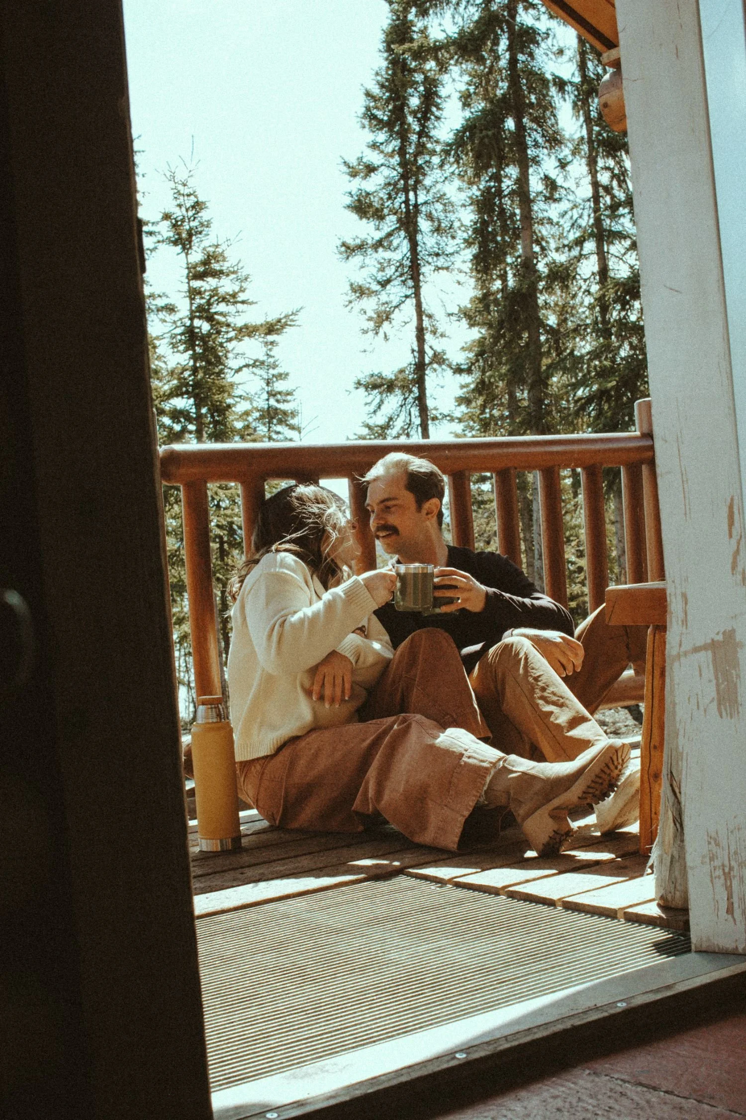 A couple sitting on a porch, smiling and holding mugs, with pine trees in the background.