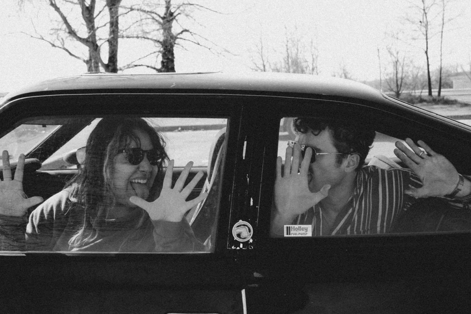 Two people inside a vintage car, smiling and making faces at the camera. The person on the left is a woman with long hair and sunglasses, sticking out her tongue. The person on the right is a man with curly hair and glasses, holding his hand to his face as if whispering or calling.