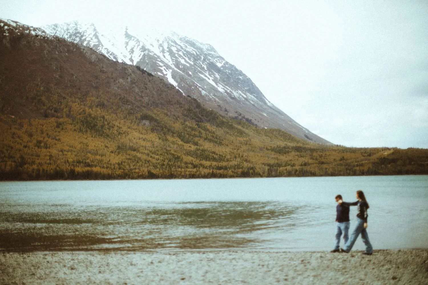 A woman and a young boy walking along the sandy shore of a lake with mountain and forest in the background.