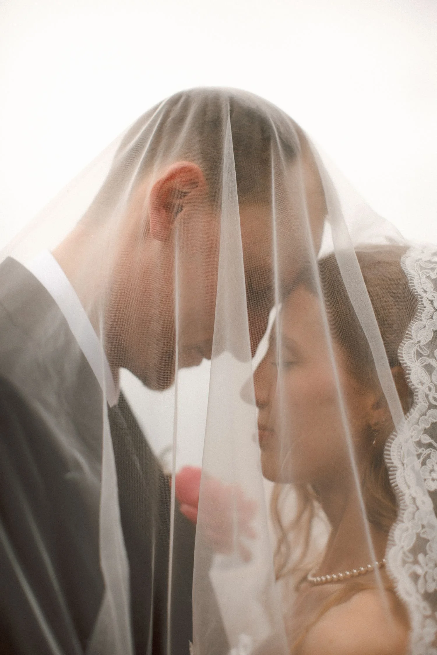 A bride and groom are behind a semi-transparent veil, with the bride wearing lace and pearls and the groom in a black suit. They are close together, with their heads inclined towards each other, in a soft, romantic setting.