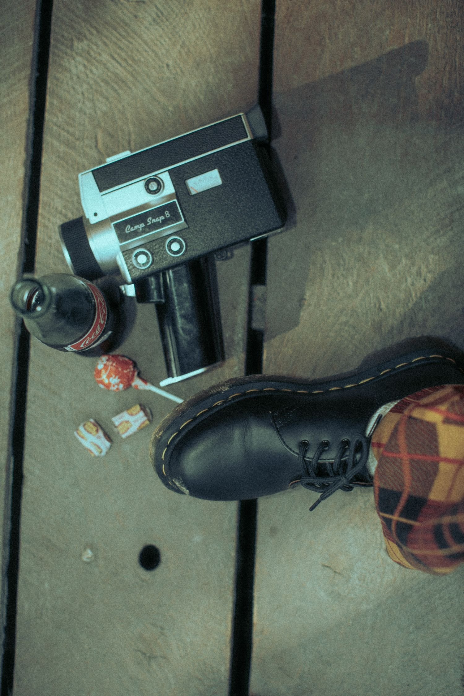 The photo shows a person's foot wearing a black boot with red plaid socks, a black metal bed frame, a wooden surface, a vintage camera, a Coca-Cola bottle, a lollipop, and some snack wrappers on a beige carpeted floor.