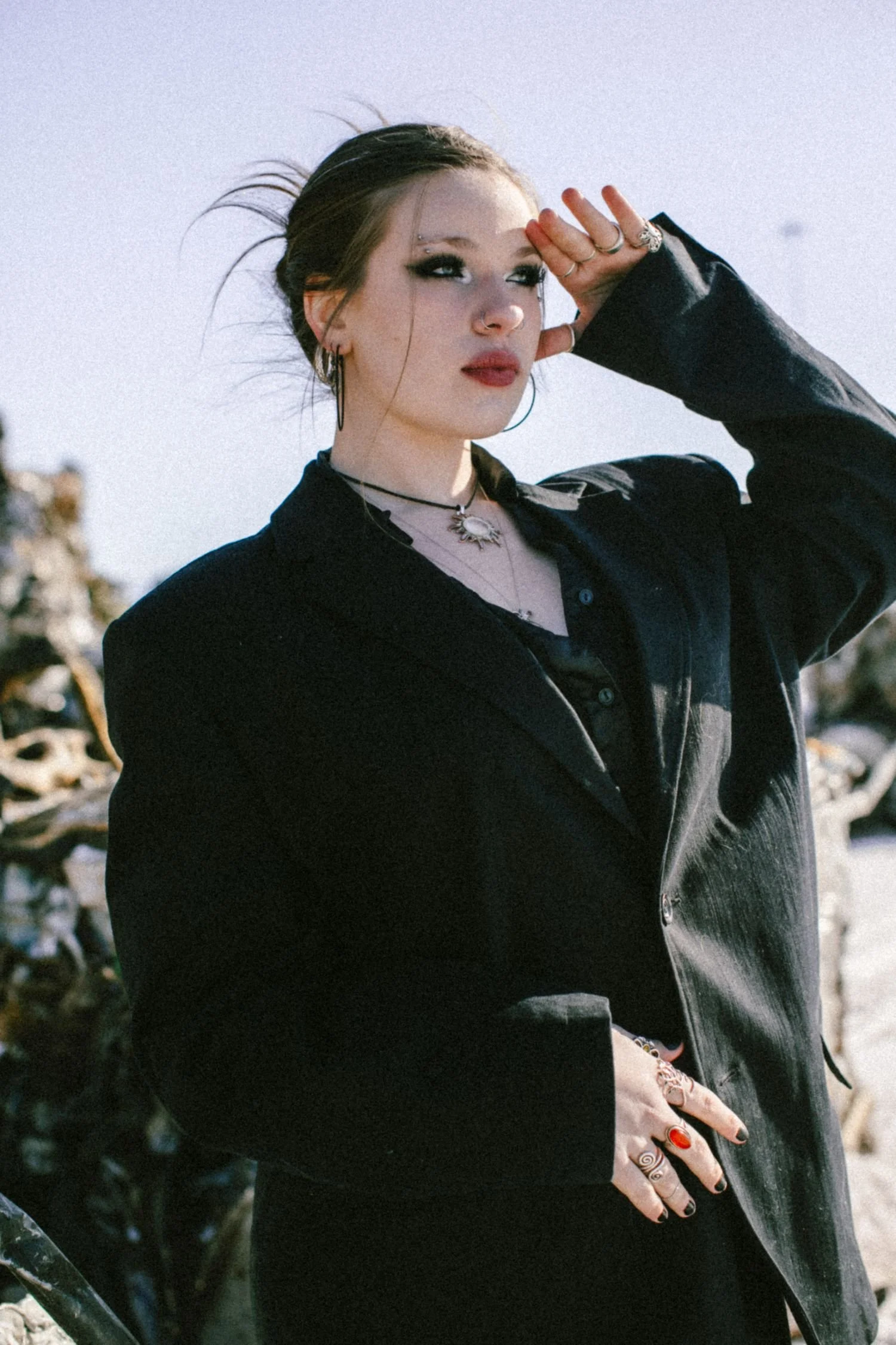 A woman with dark hair, multiple rings, and eye makeup, wearing a black jacket and making a saluting gesture with her right hand, outdoors near rocks and trees.