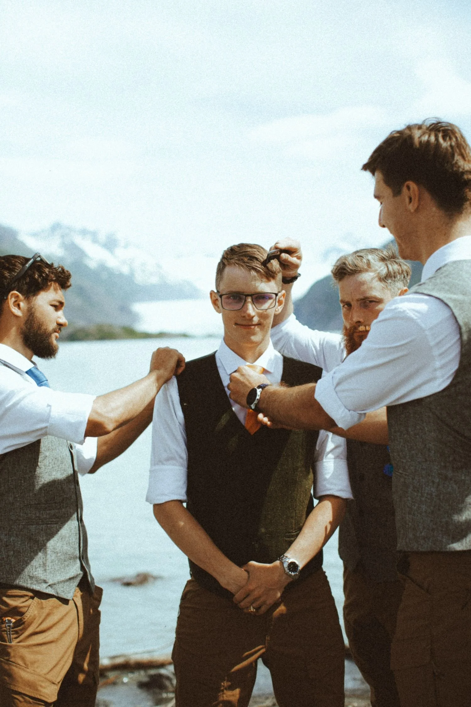 A group of five men standing outdoors by a body of water, with mountains in the background. They are dressed in semi-formal attire and appear to be adjusting their clothes and styling their hair.