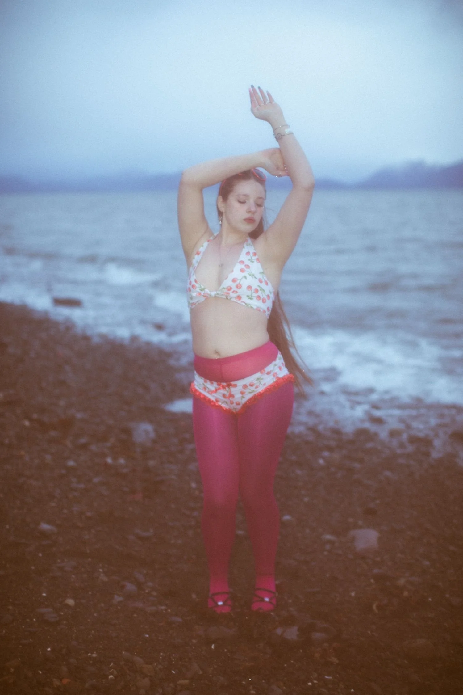 A woman wearing a floral bikini top, pink leggings, and high heels standing on a rocky beach by the ocean with eyes closed and arms raised above her head.