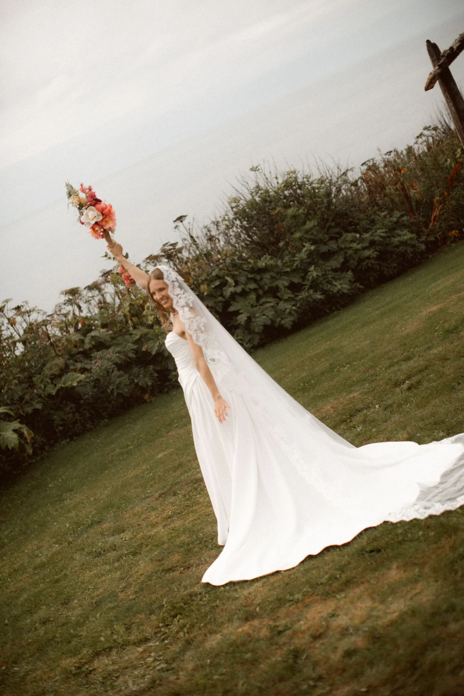 A bride in a wedding dress holding a bouquet of flowers, smiling, outdoors on a grassy area with bushes and trees in the background.