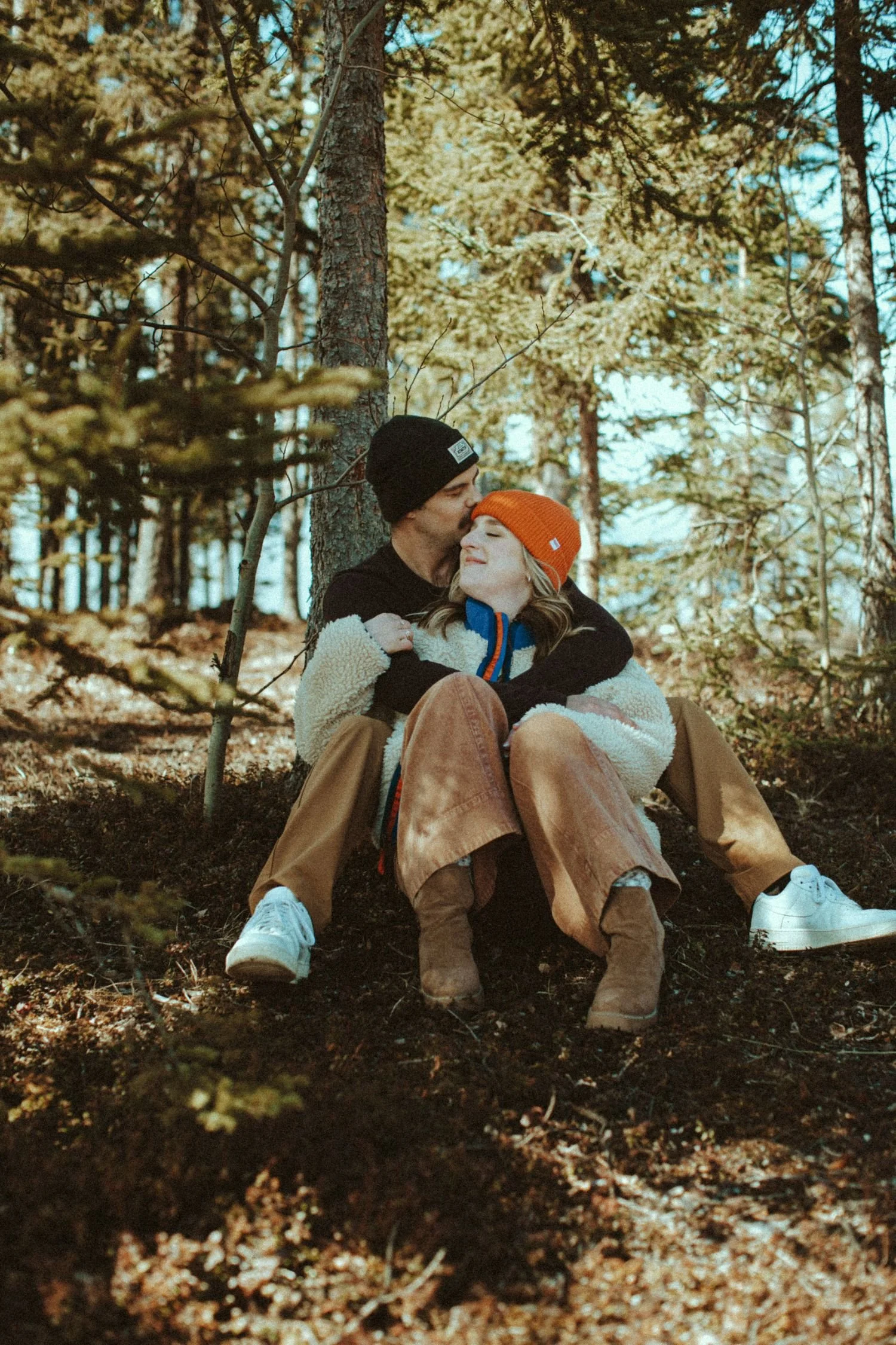 A couple sitting in a wooded area, hugging and smiling with trees in the background.