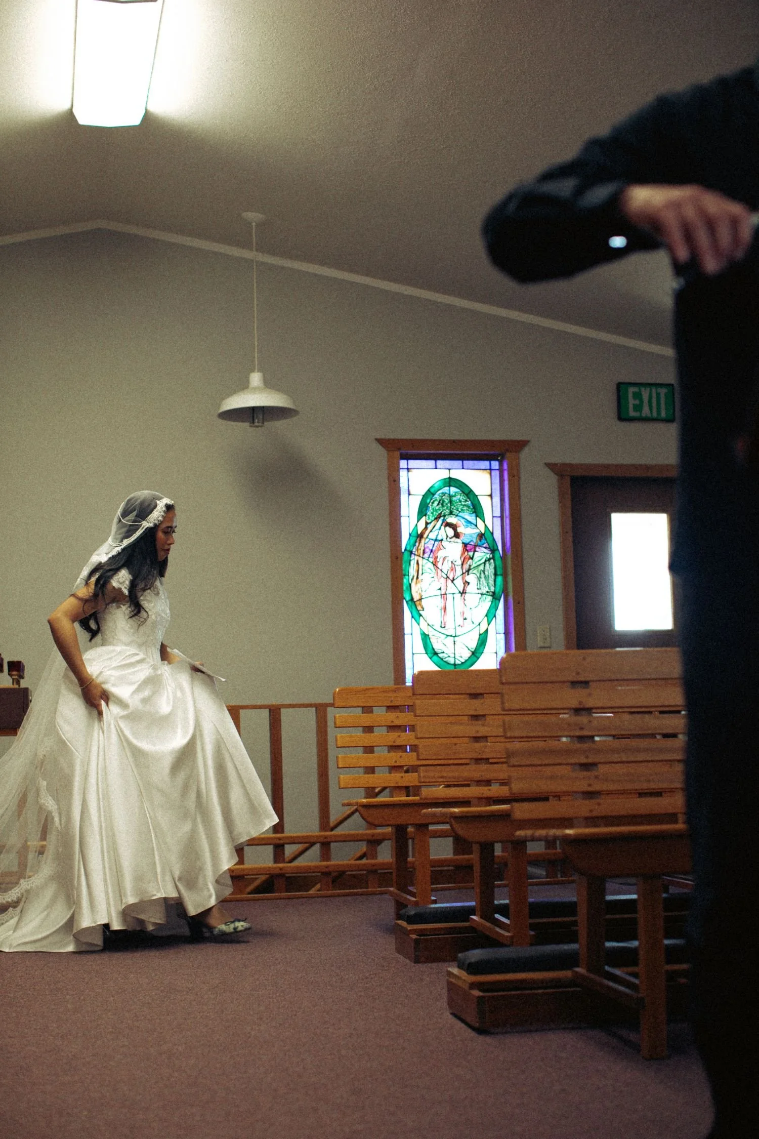 A woman in a white wedding dress and veil standing in a church with wooden pews, stained glass window, and hanging light fixtures.