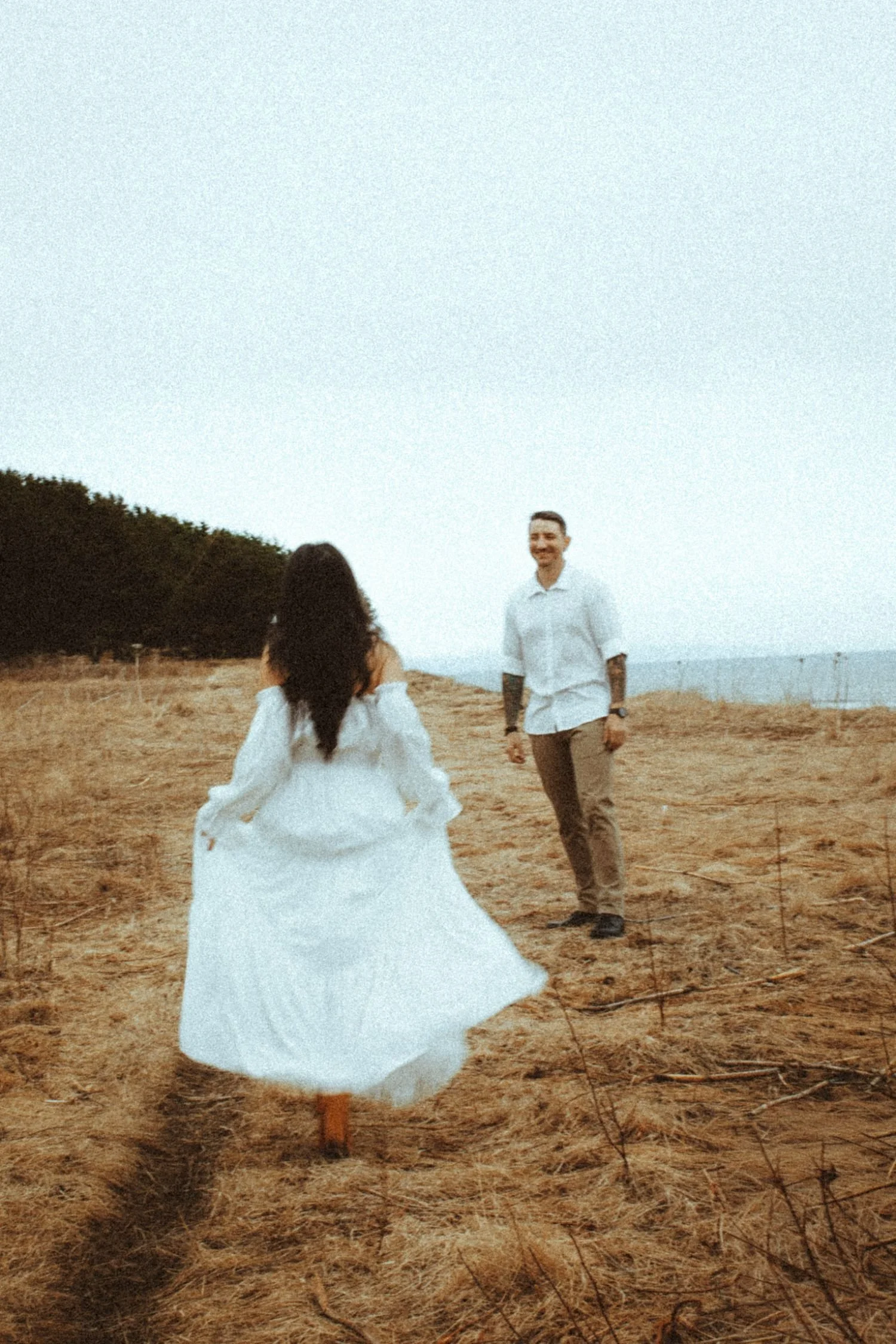 A woman in a white dress running in a field with dry grass, with a man in a white shirt and khaki pants walking towards her, under a clear sky with trees in the background.