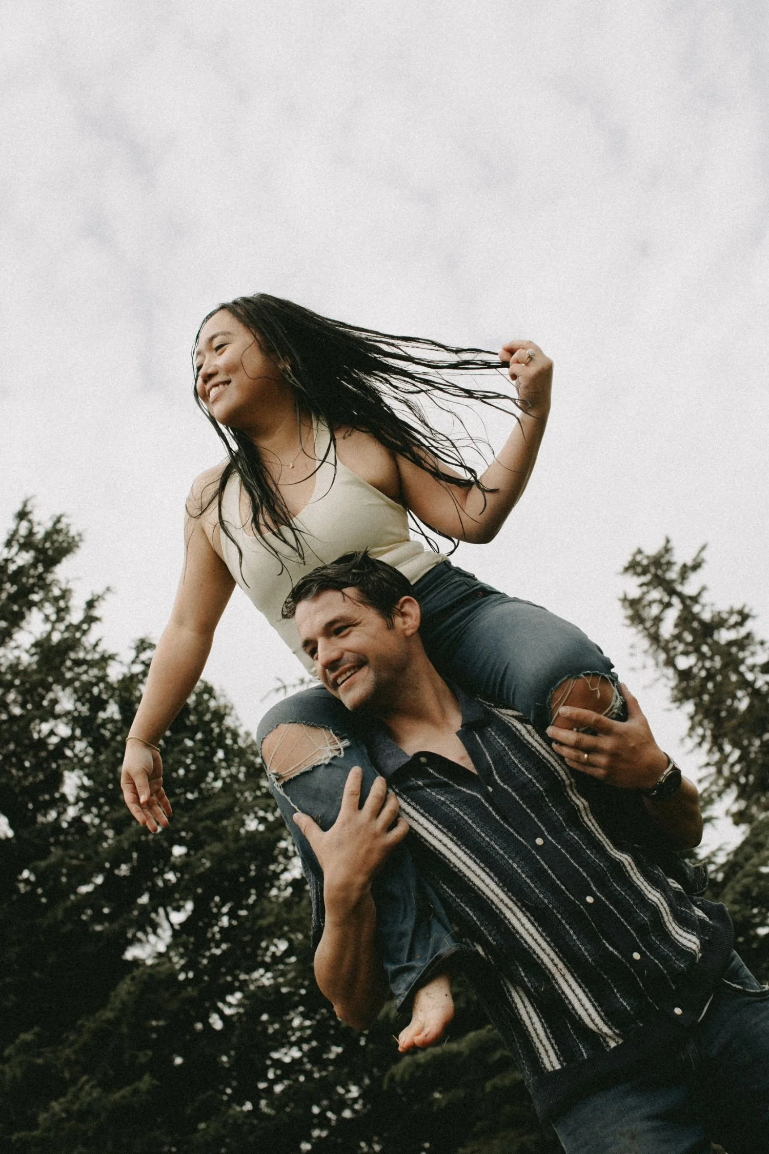 A young woman with long black hair, smiling and playing, is sitting on the shoulders of a man with dark hair, who is also smiling. They are outdoors with trees and an overcast sky in the background.
