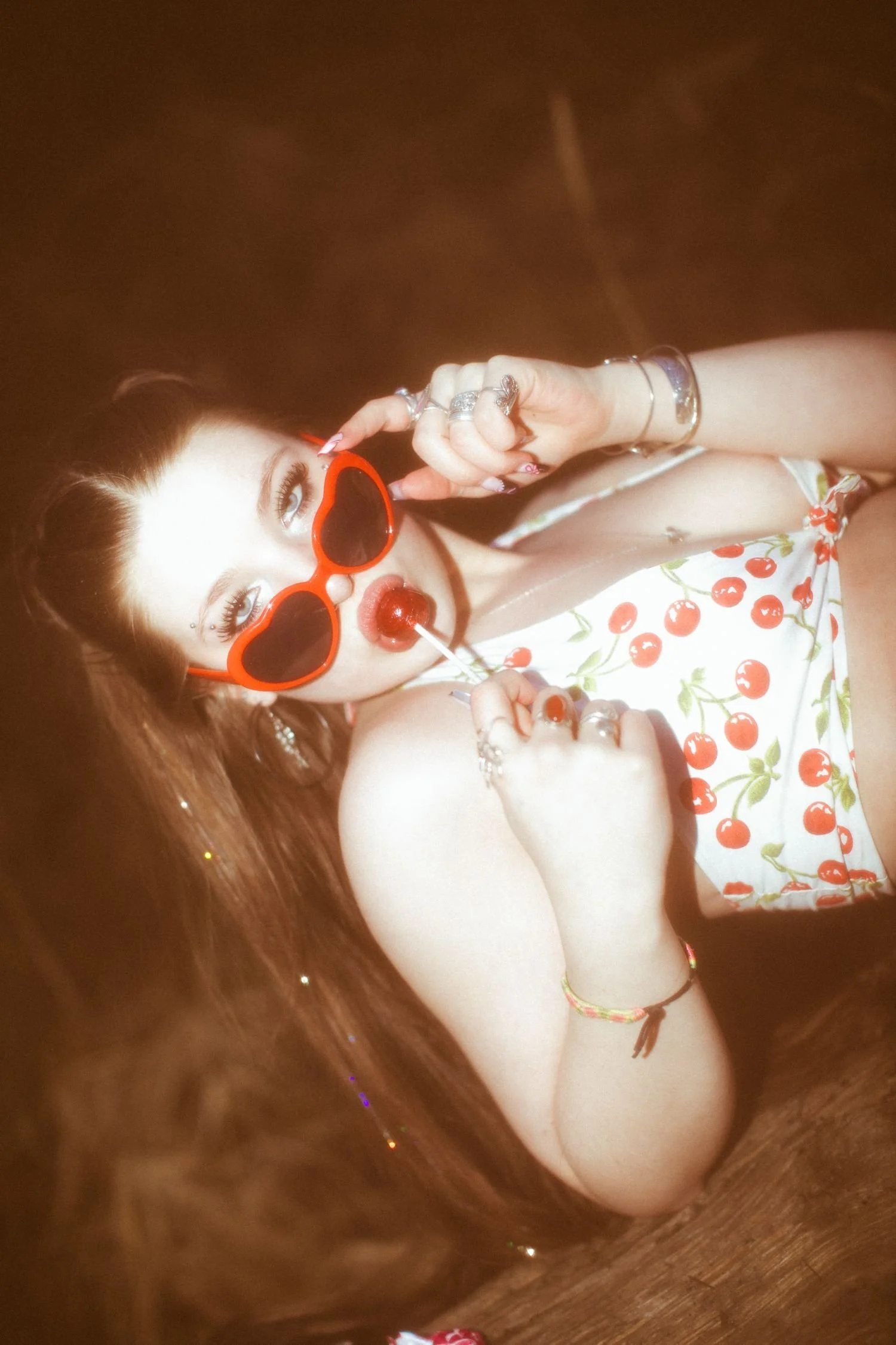 A young woman with long red hair, wearing heart-shaped sunglasses and a cherry-patterned dress, is lying on the floor. She is holding a lollipop near her lips and looking into the camera.