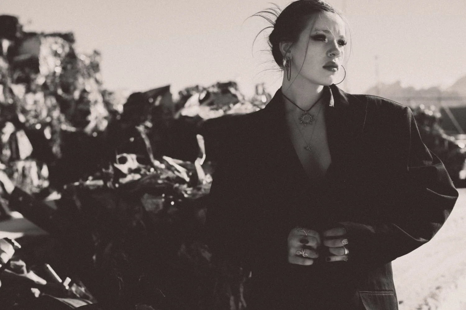 Editorial fashion photo of a woman with styled hair, makeup, hoop earrings, and jewelry, wearing a dark jacket, standing outdoors next to a pile of rocks or rubble.