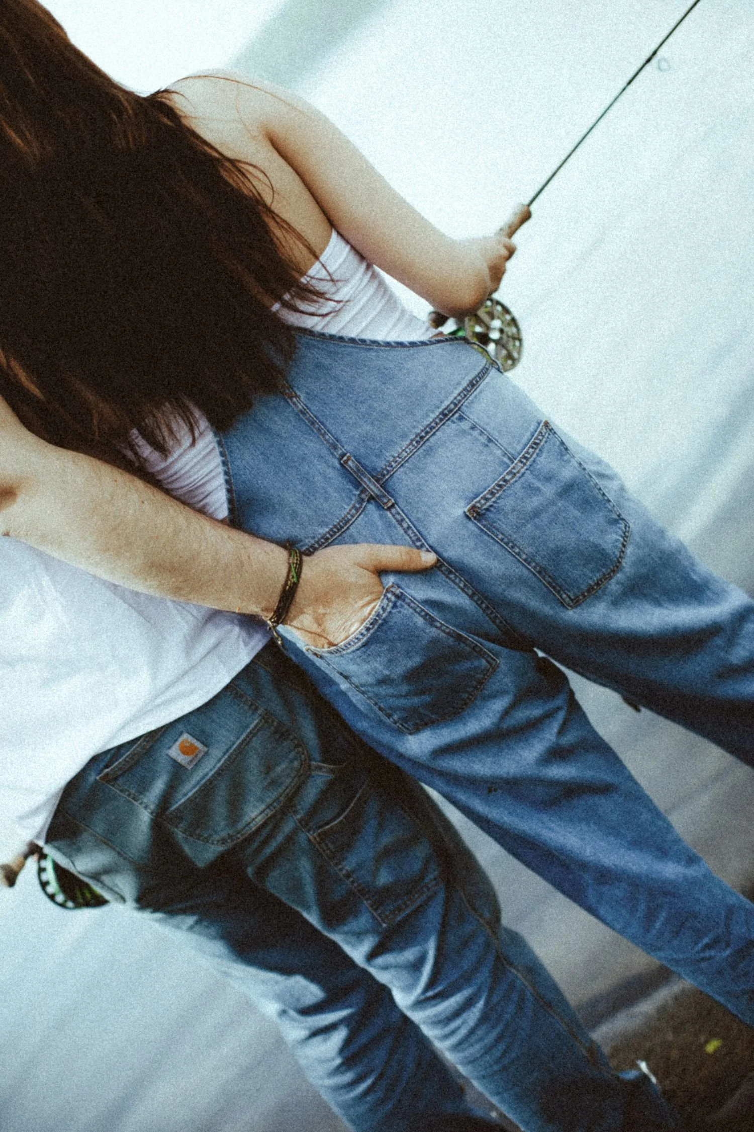 A couple, the woman has long, dark hair wearing a white tank top and blue denim overalls, holding a fishing rod while standing on a concrete surface.
