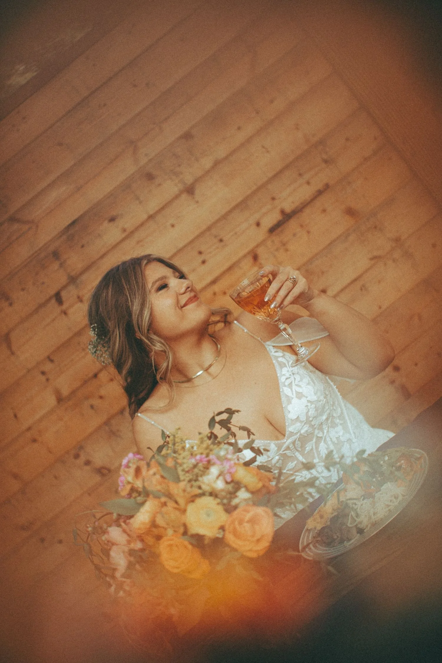 A woman in a white lace wedding dress holding a glass of wine, smiling, with a bouquet of flowers in the foreground and a wooden wall in the background.