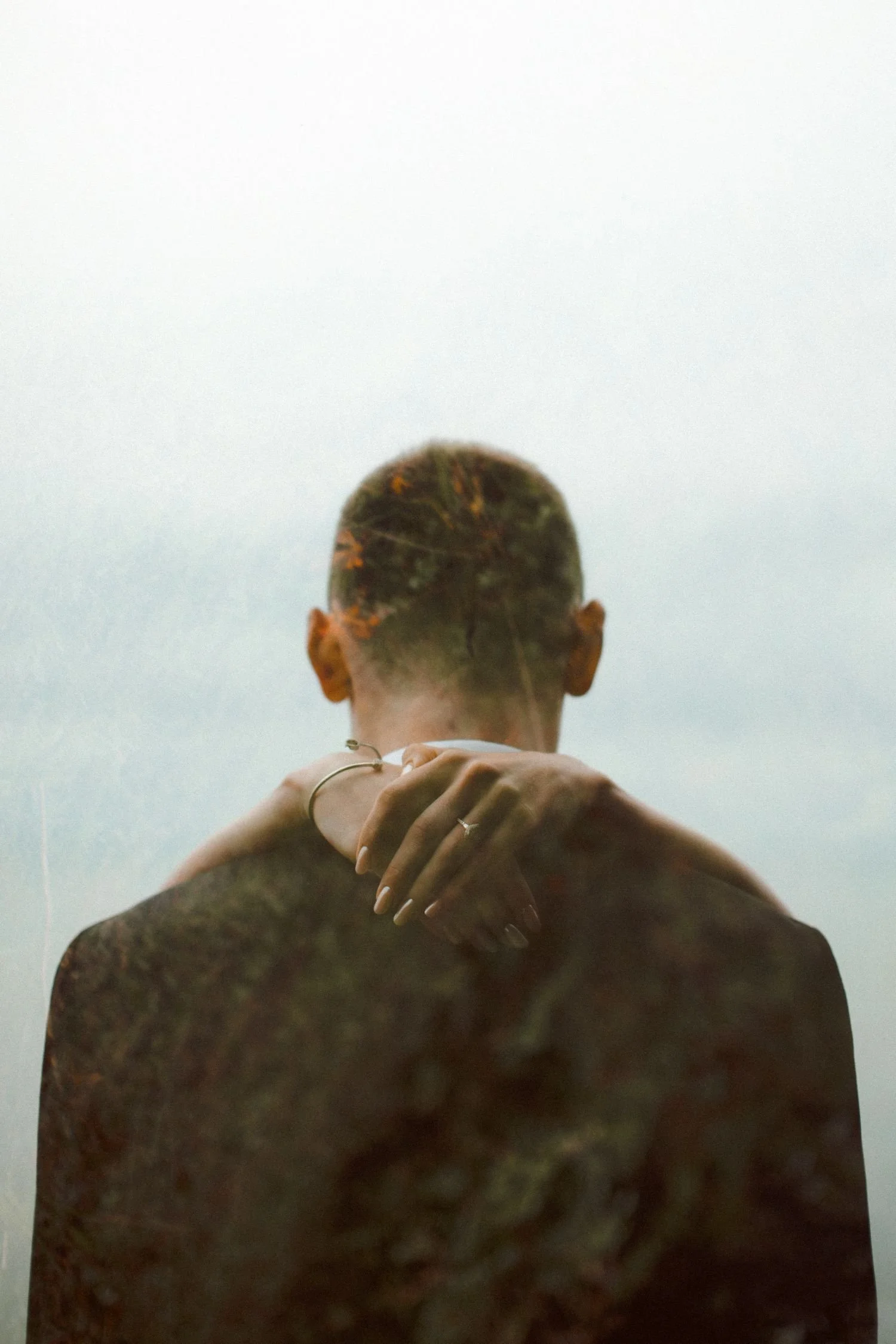 A person with tattoos on their back and a bun hairstyle is standing with their back to the camera. They are holding another person's hand and shoulder gently, with a minimal background.