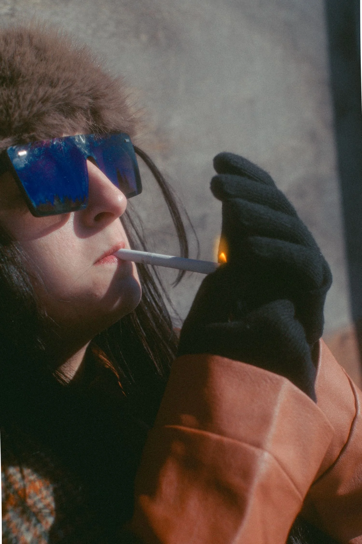 A woman wearing reflective sunglasses with a blue and purple tint, smoking a cigarette. She has brown hair and is wearing black gloves and a brown and beige coat.