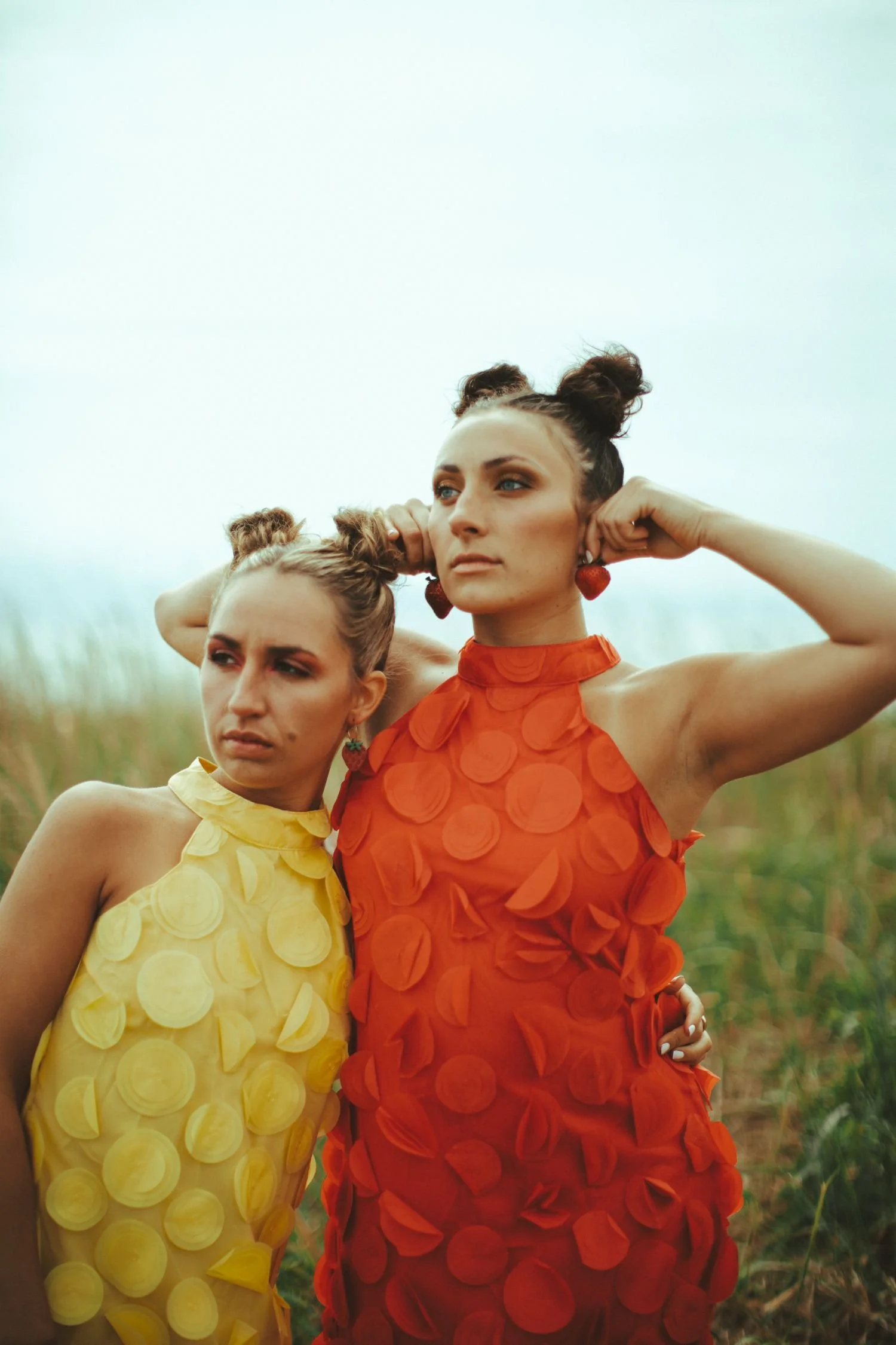 Two women standing outdoors in a field with tall grasses, wearing bright, colorful dresses with circular patterns. One woman has on a yellow dress, and the other on a red dress. Both women have their hair styled in space buns and are wearing earrings. The woman in red has earrings that look like strawberries and is crossing her arms, while the other woman is looking to the side.