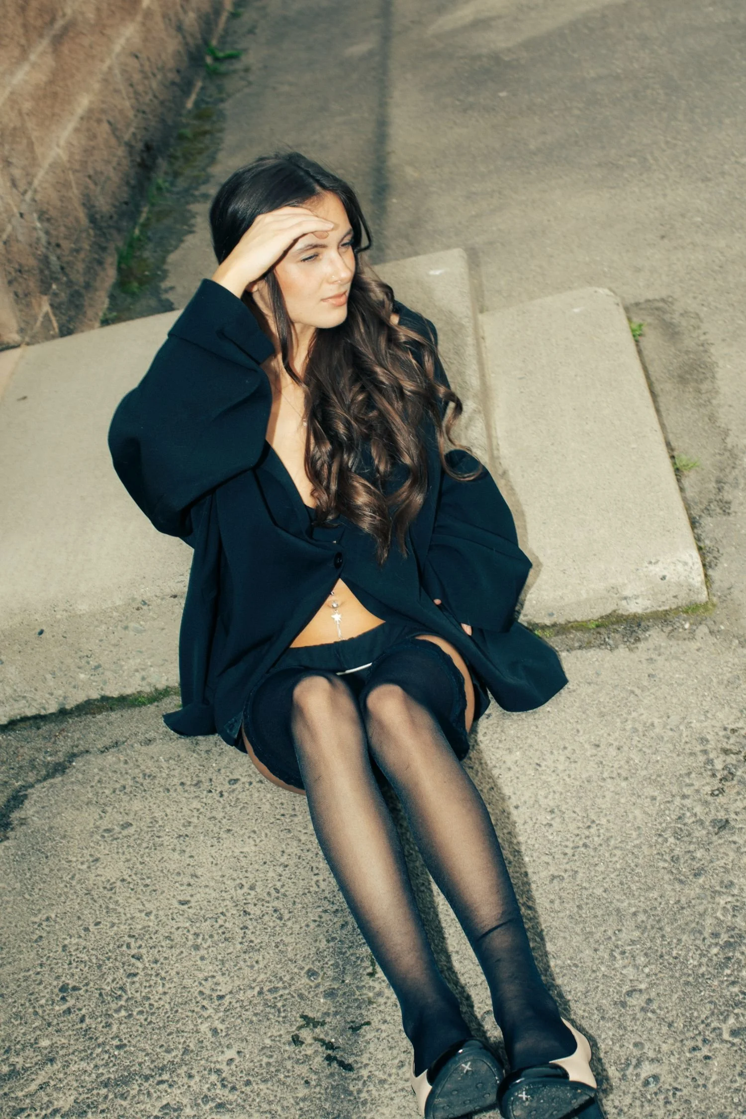 A woman sitting on the ground against a concrete wall, wearing a black blazer, black stockings, and black shoes, with long dark hair and holding her forehead while looking away.
