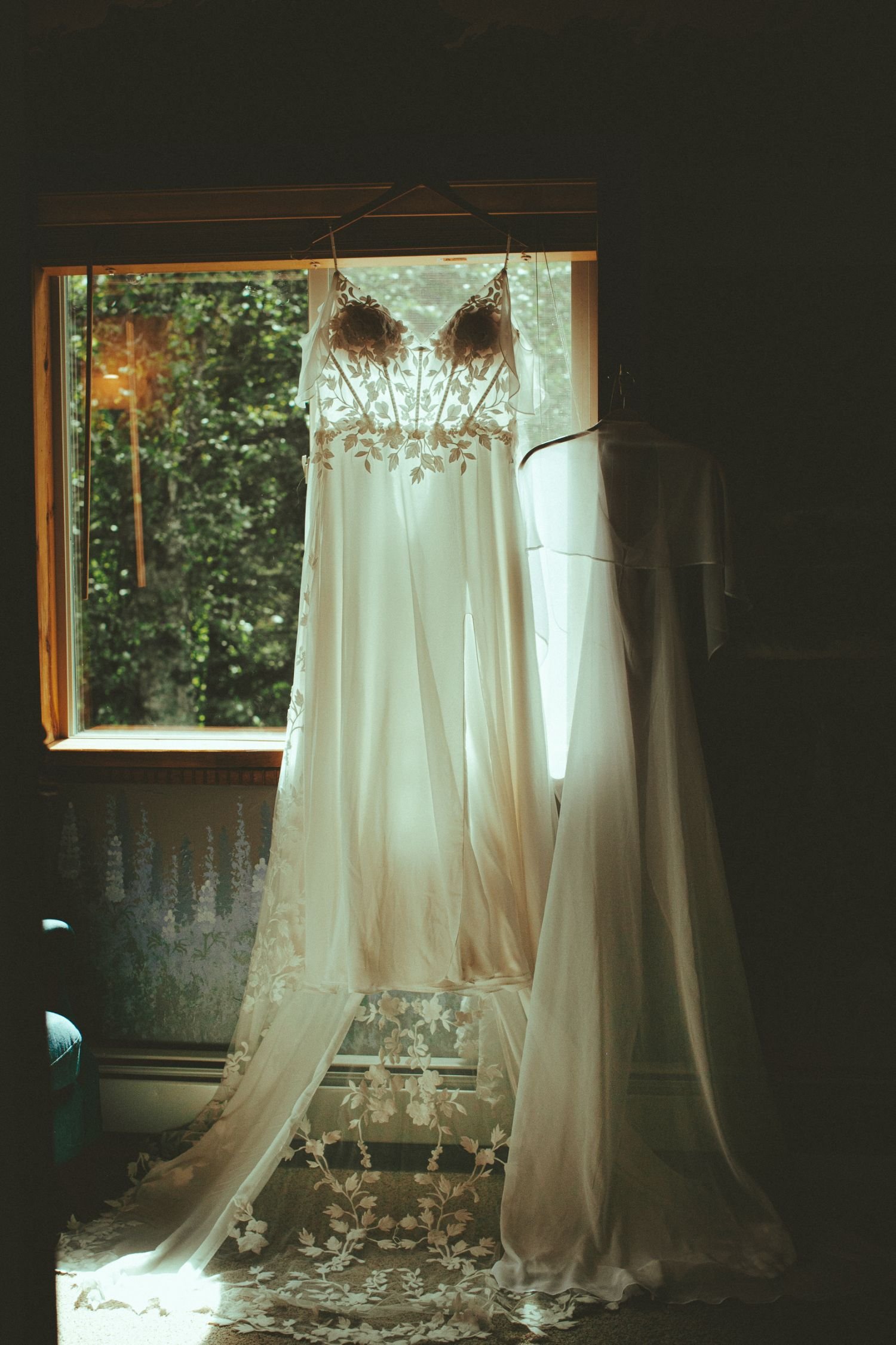 A white wedding dress with floral embroidery hanging in front of a window with sunlight coming through, with a sheer lace curtain partially draped on the floor.