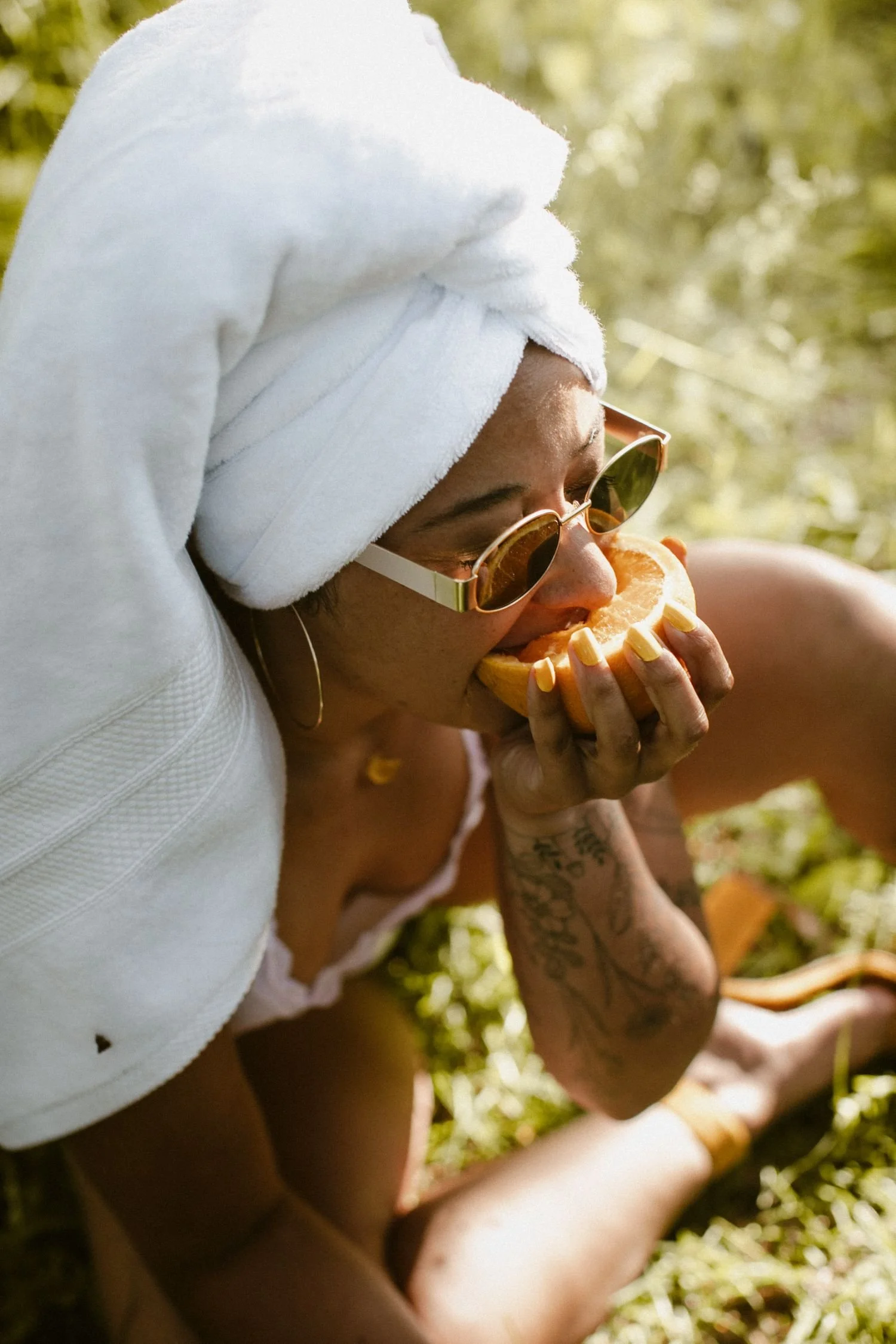 A woman with a white towel wrapped around her head, wearing sunglasses, is eating a slice of melon outdoors. She has a tattoo on her arm and is sitting in a natural, sunny environment.