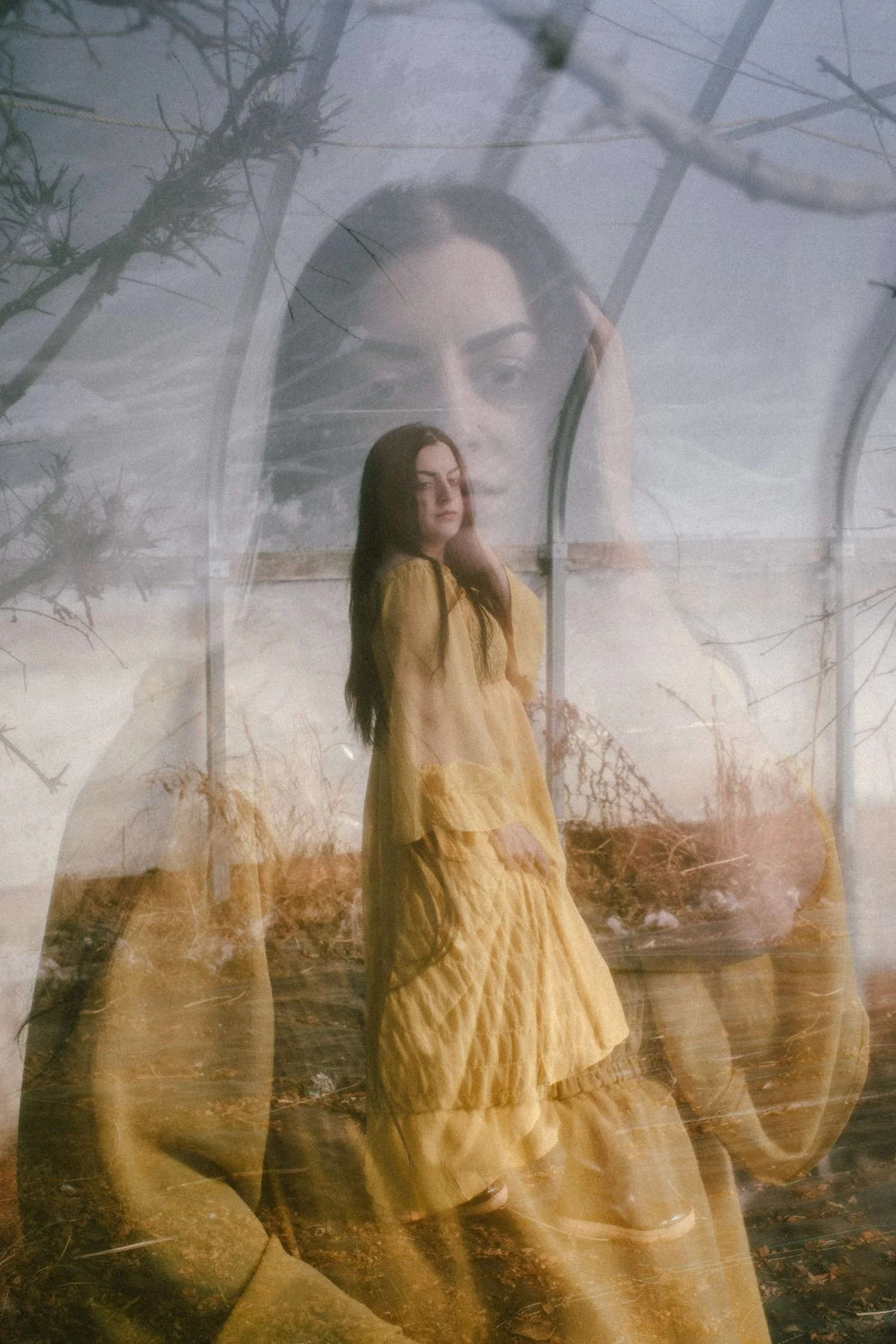 A woman with long dark hair wearing a yellow dress standing outdoors, with trees and a cloudy sky in the background, and an overlaid second, larger, semi-transparent image of her face.