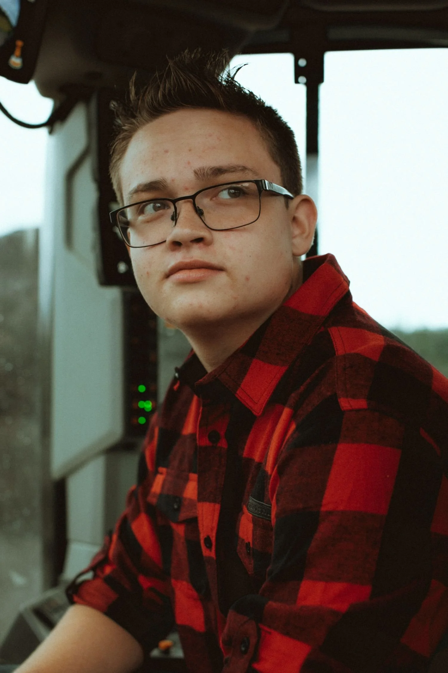 A young man with short dark hair, glasses, wearing a red and black plaid shirt, sitting at a computer or gaming console.