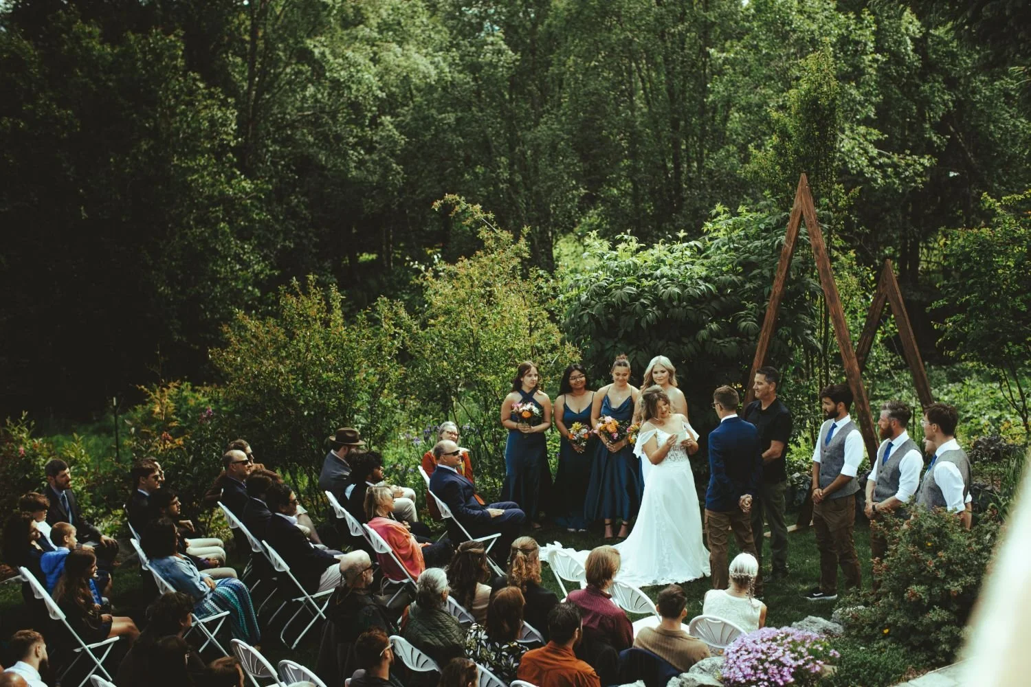 A wedding ceremony taking place outdoors in a lush garden with trees and greenery. The bride and groom are standing with bridesmaids and groomsmen, surrounded by seated guests.