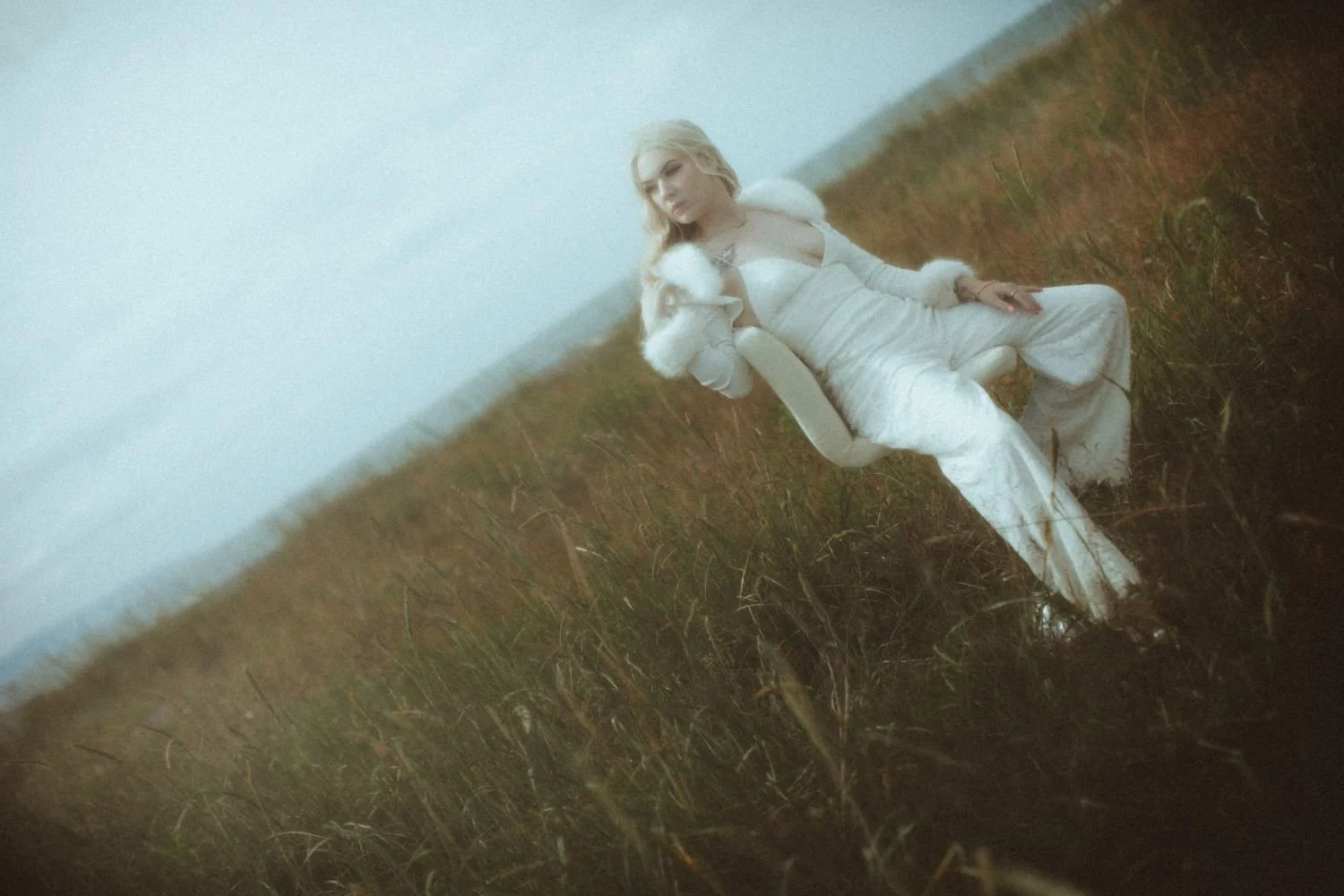 A woman in a white dress and fur coat sitting on the grass in a field, with a cloudy sky behind her.
