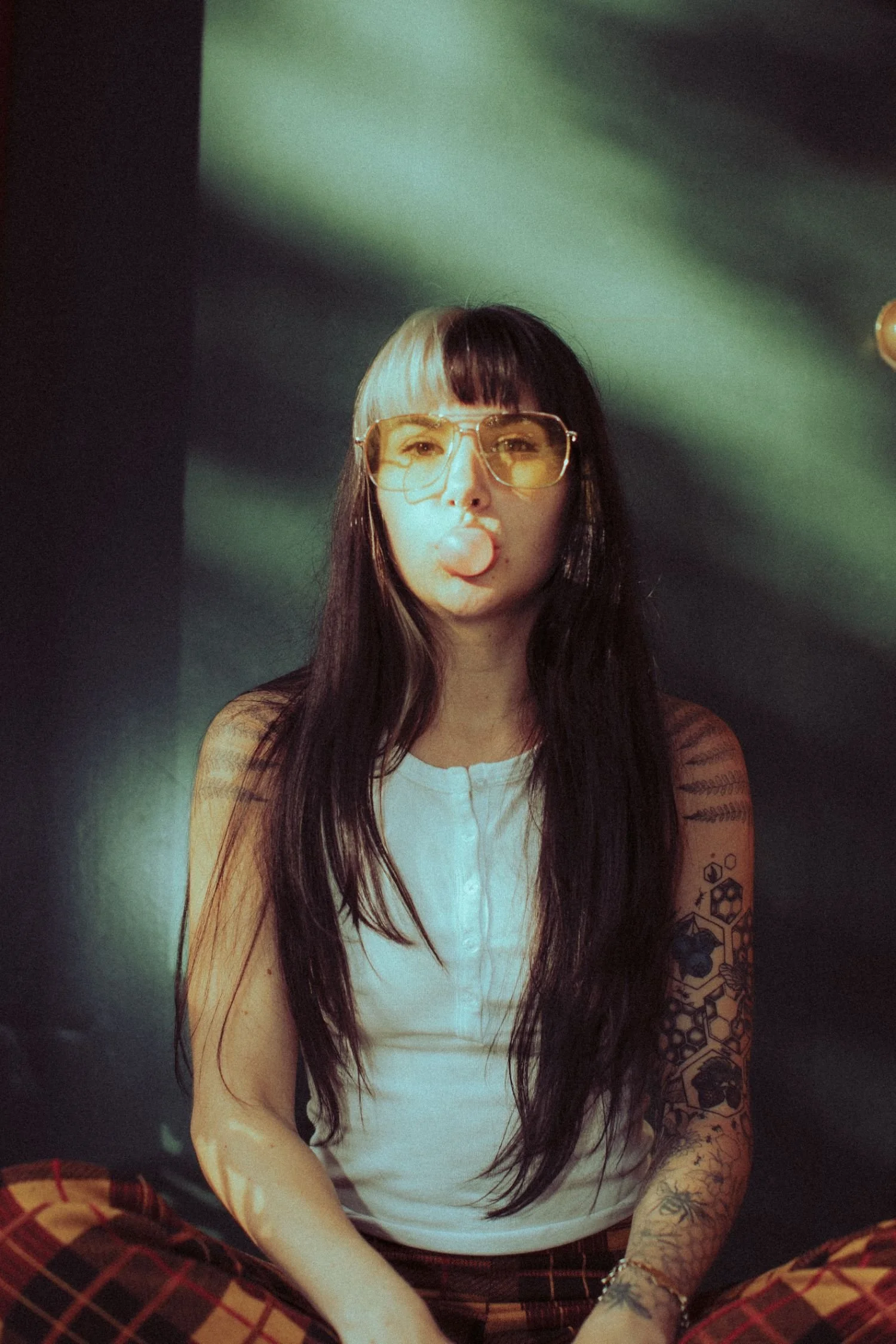 Danika Stiles with long dark hair, wearing sunglasses and a white sleeveless shirt, is sitting cross-legged, blowing a bubble gum bubble. She has tattoos on her right arm and a tattoo on her left forearm.