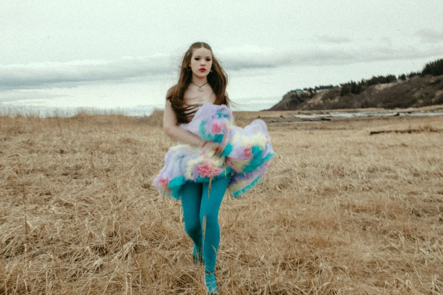 Young woman walking through a grassy field holding a colorful pastel blanket, with a cloudy sky and distant hills in the background.