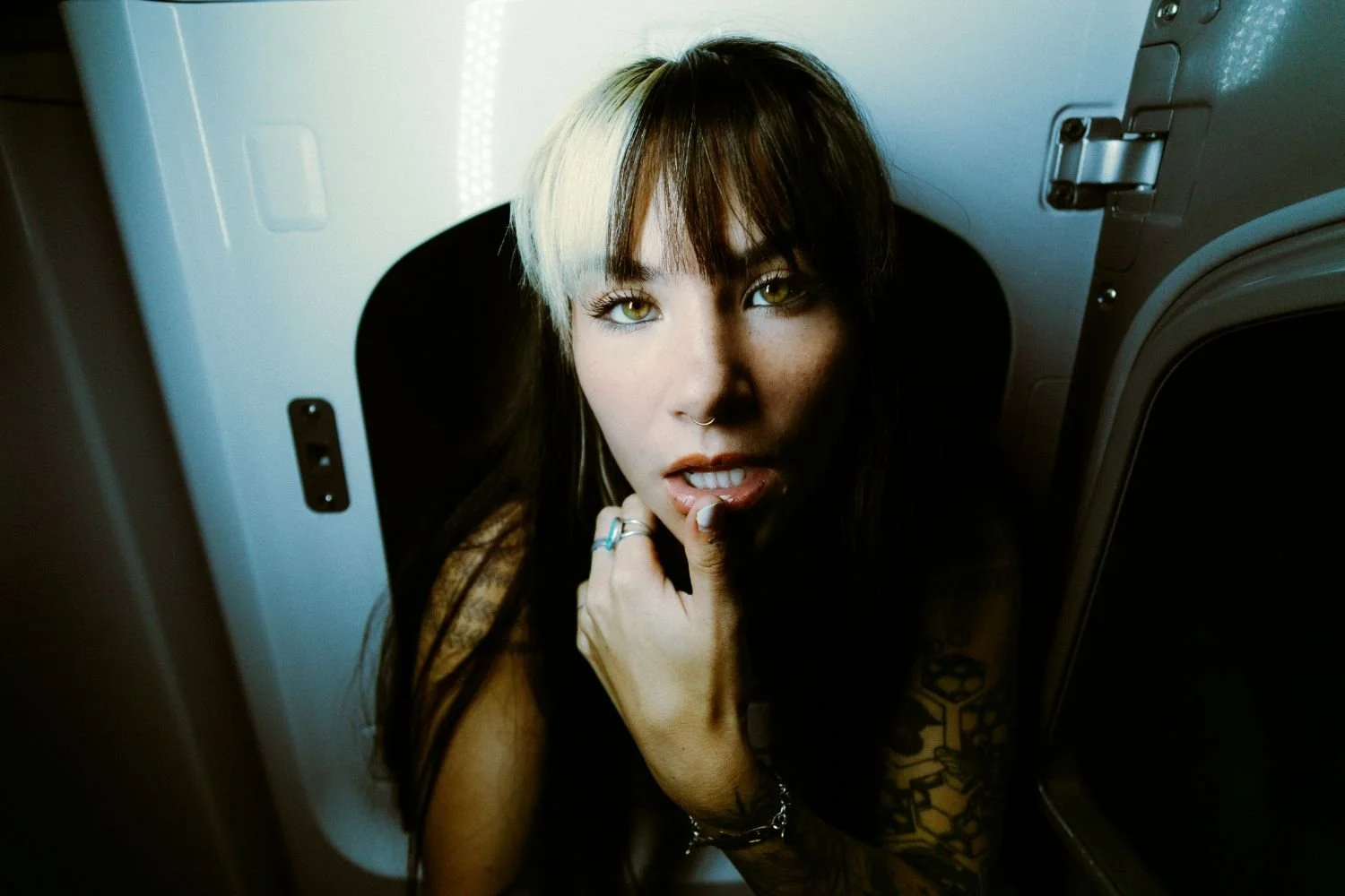 Danika Stiles with long dark hair, blonde streaks, tattoos, wearing rings and a bracelet, looking directly at the camera with her finger on her lips, inside a laundry dryer or appliance.