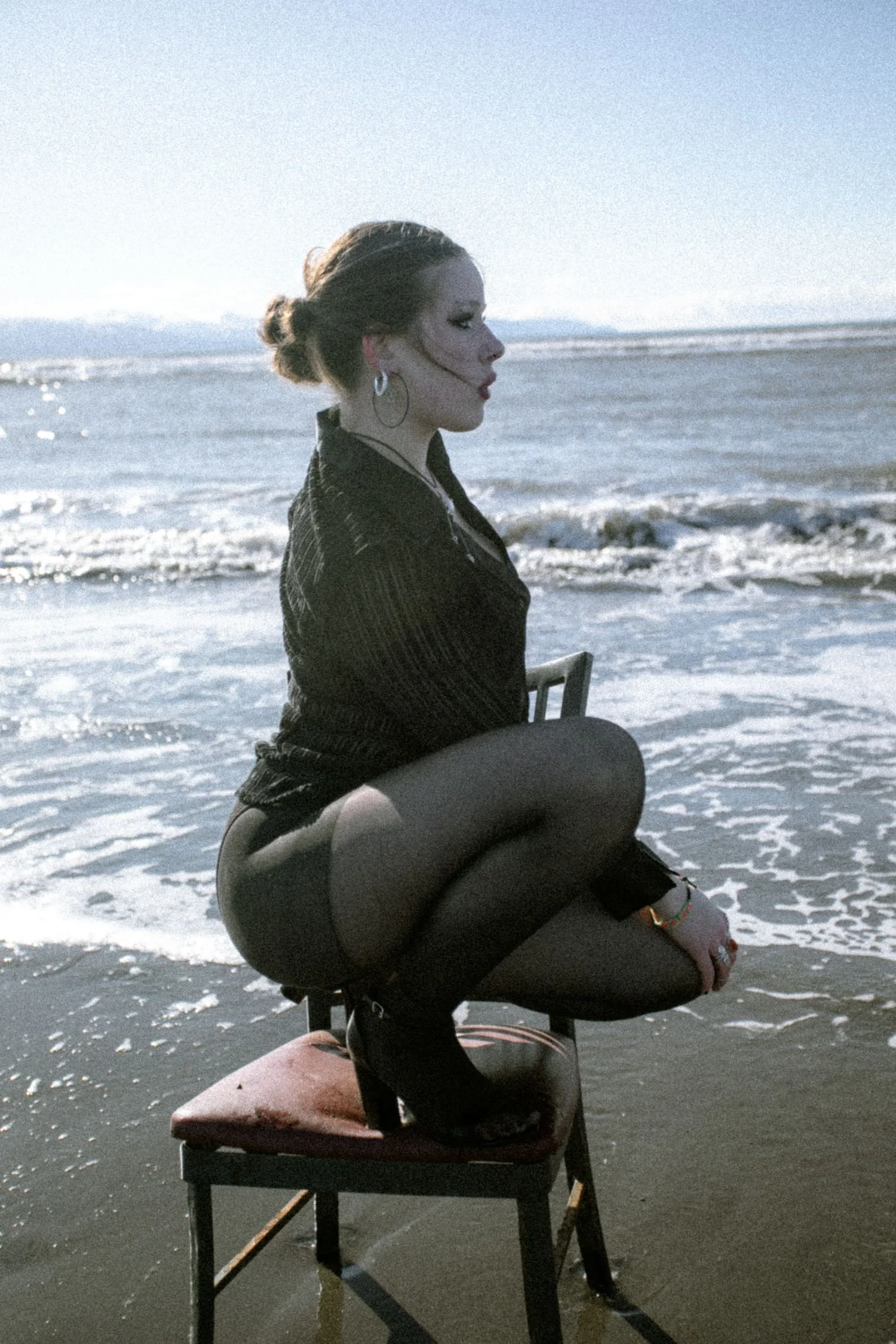 A woman with her hair in a bun sitting on a chair at the beach, facing to the right, with the ocean and waves in the background, wearing hoop earrings and a dark striped jacket.