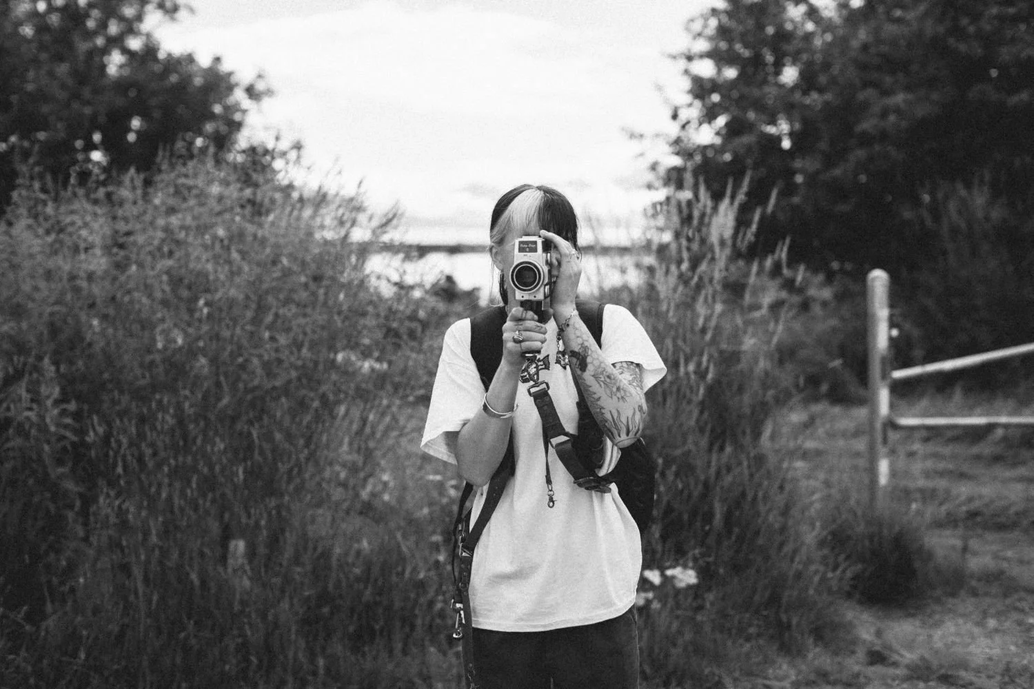 Danika Stiles taking a video with a super 8 camera outdoors in a natural setting with tall grasses and trees, in black and white.