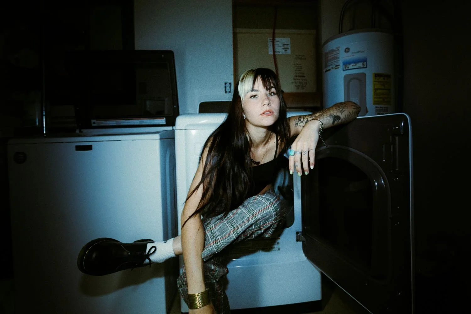 Danika Stiles with long dark hair, wearing a black top and plaid pants, crouches by a washing machine in a dark laundry room, looking at the camera.
