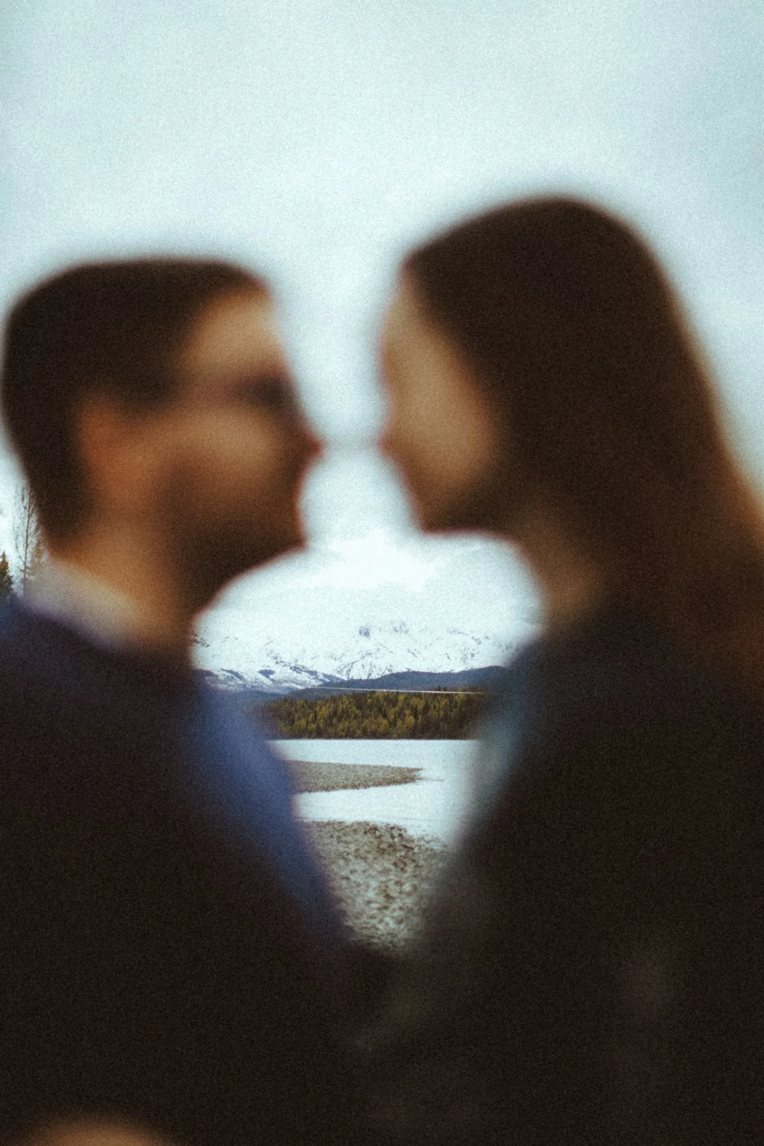 A blurred photo of two people face to face, with mountains, trees, and a body of water in the background, seen through a heart-shaped opening in an object, possibly a stone or a shell.