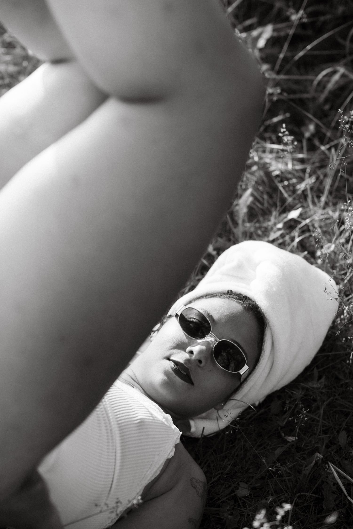 A woman lying on her back in grass, wearing sunglasses and a head wrap, with her arm extended towards the camera.