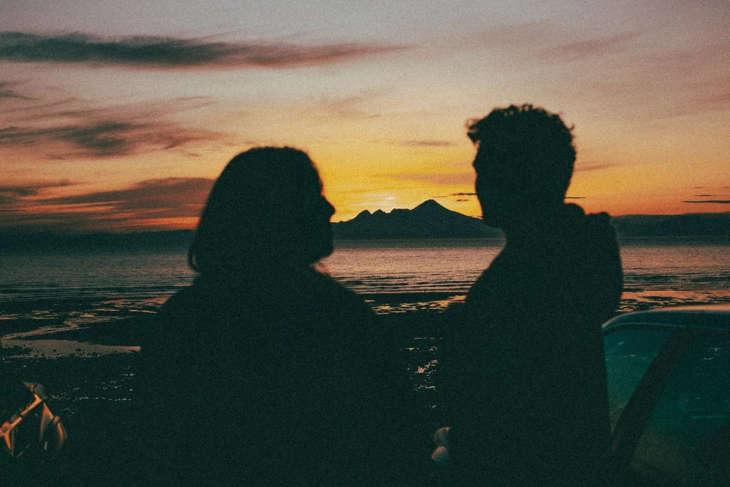 Silhouettes of a man and woman facing each other at sunset by a lake, with mountains in the background.