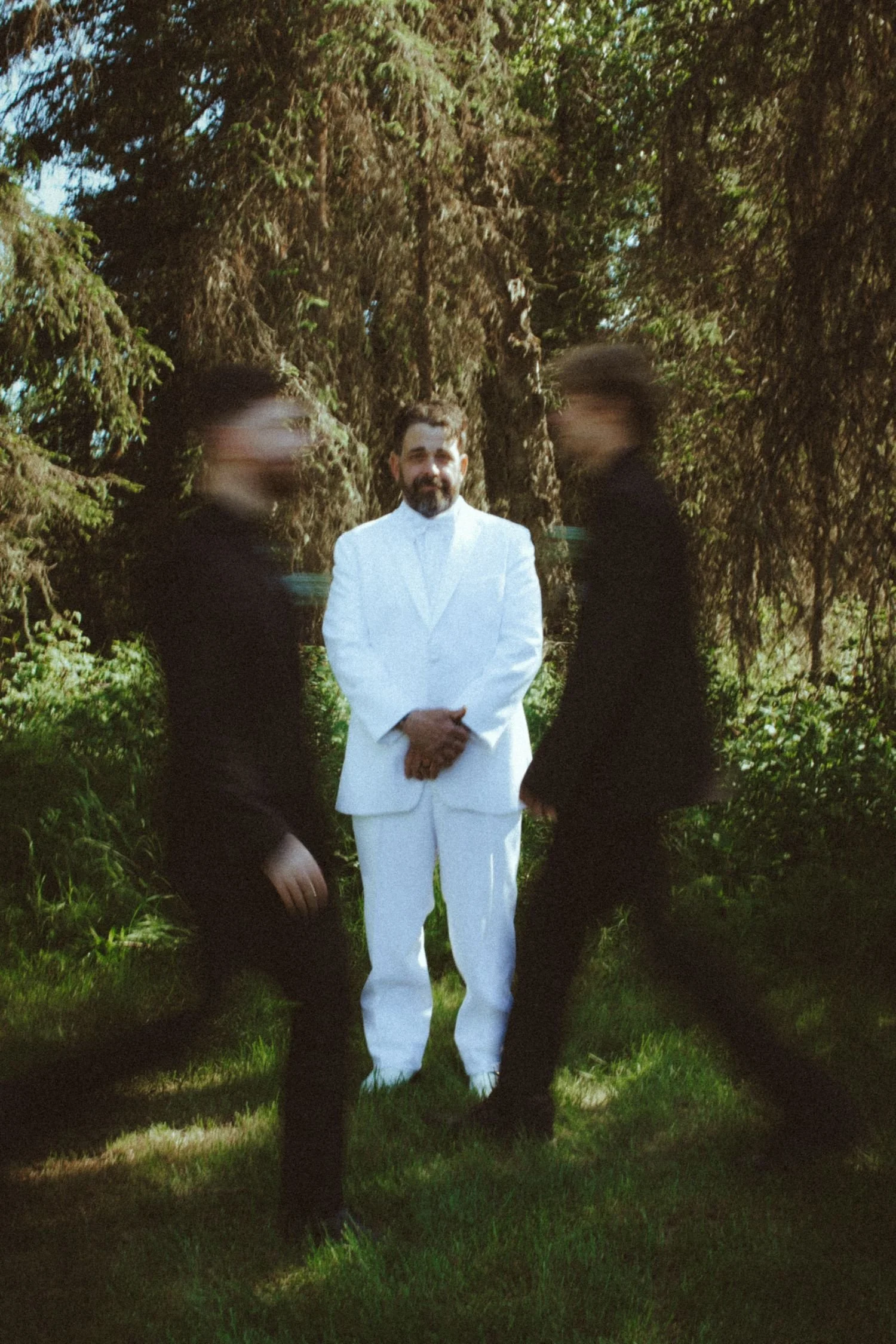 A man in a white suit and two people in black clothing standing outdoors among green trees and grass, with one person's face blurred.