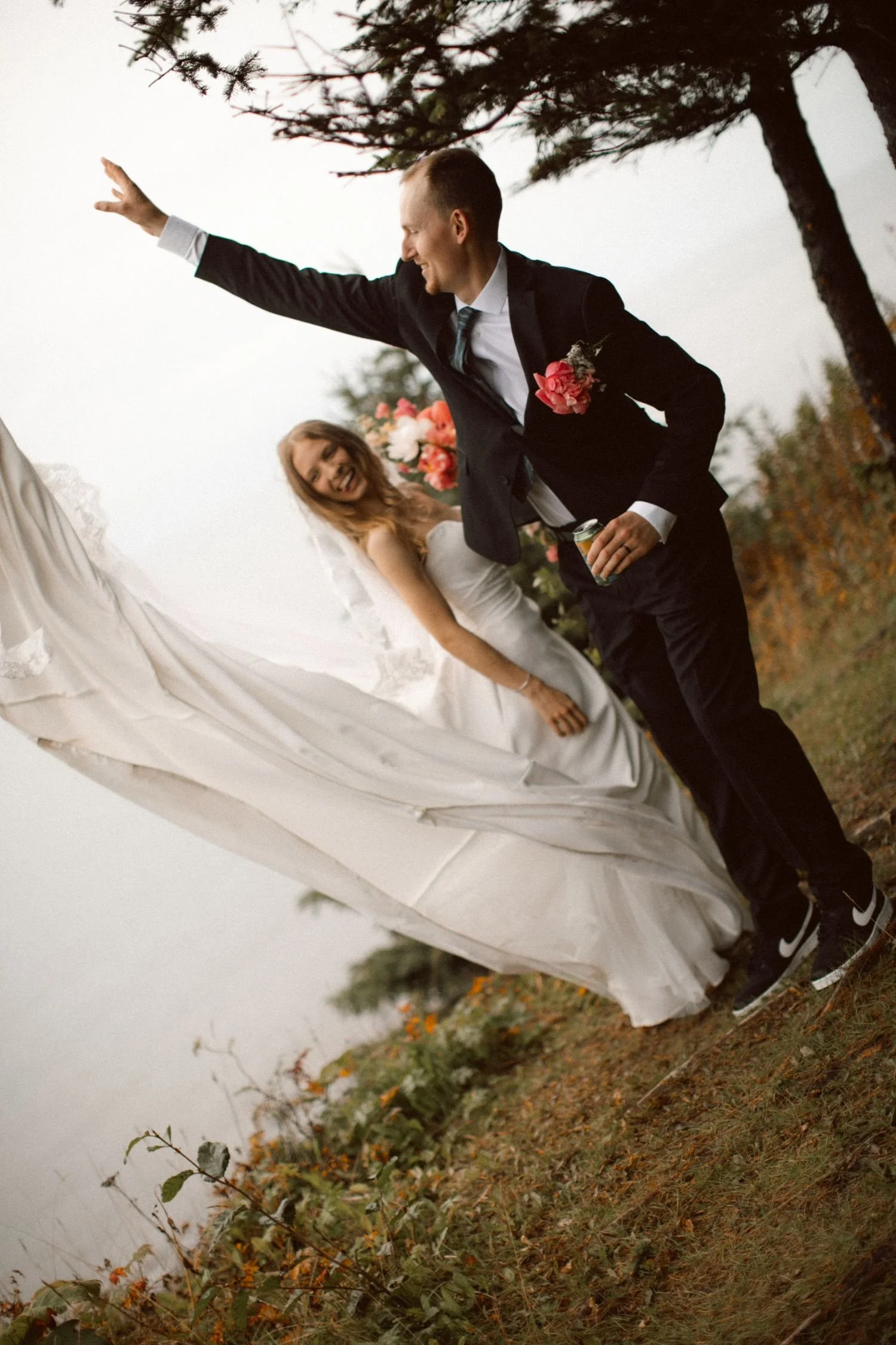 A man in a black suit and a woman in a white wedding dress at an outdoor wedding ceremony. The man is wearing sneakers and holding a beer, while the woman is smiling and looking at him. There are trees and a white draped backdrop with floral decorations behind them.